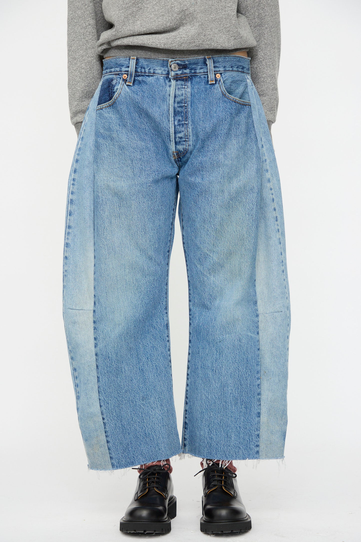 A person wears B Sides Vintage Lasso Jean in Indigo—two-tone blue with raw hems—paired with a gray sweater and black shoes, channeling vintage denim style against a plain white background.