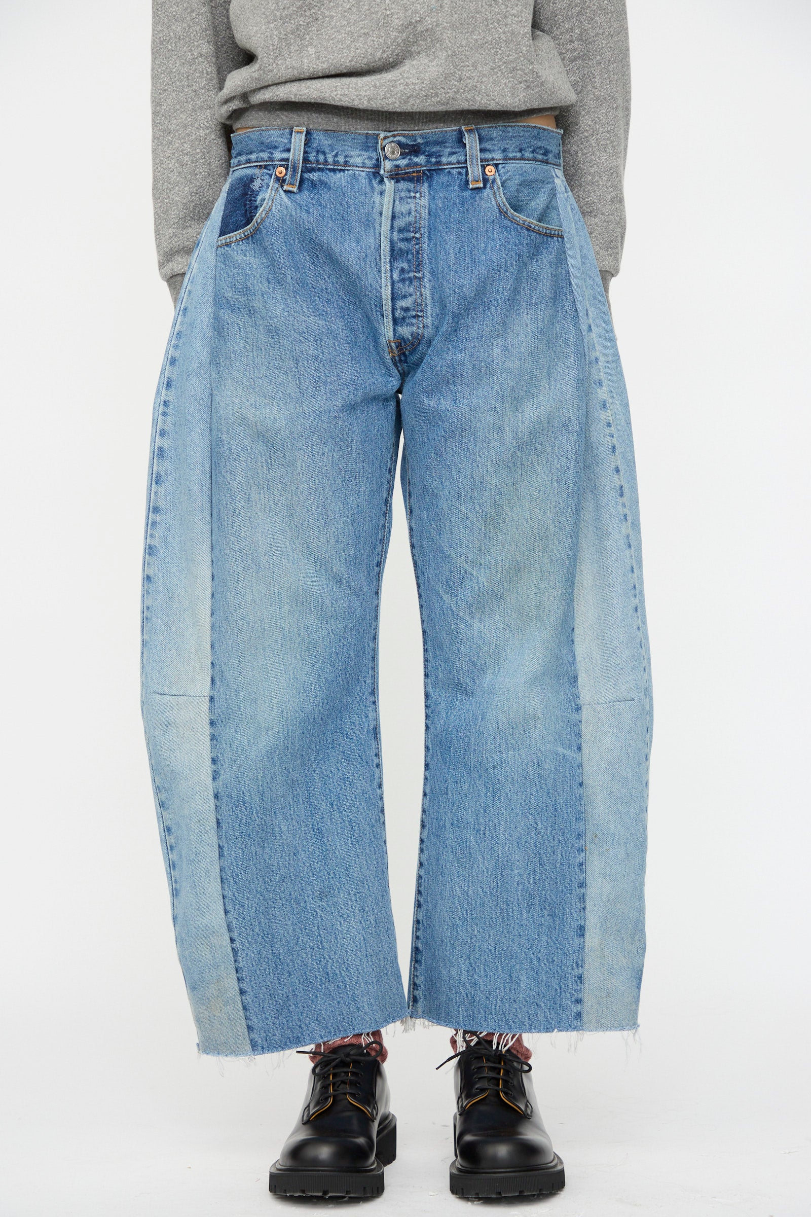 A person wears B Sides Vintage Lasso Jean in Indigo—two-tone blue with raw hems—paired with a gray sweater and black shoes, channeling vintage denim style against a plain white background.
