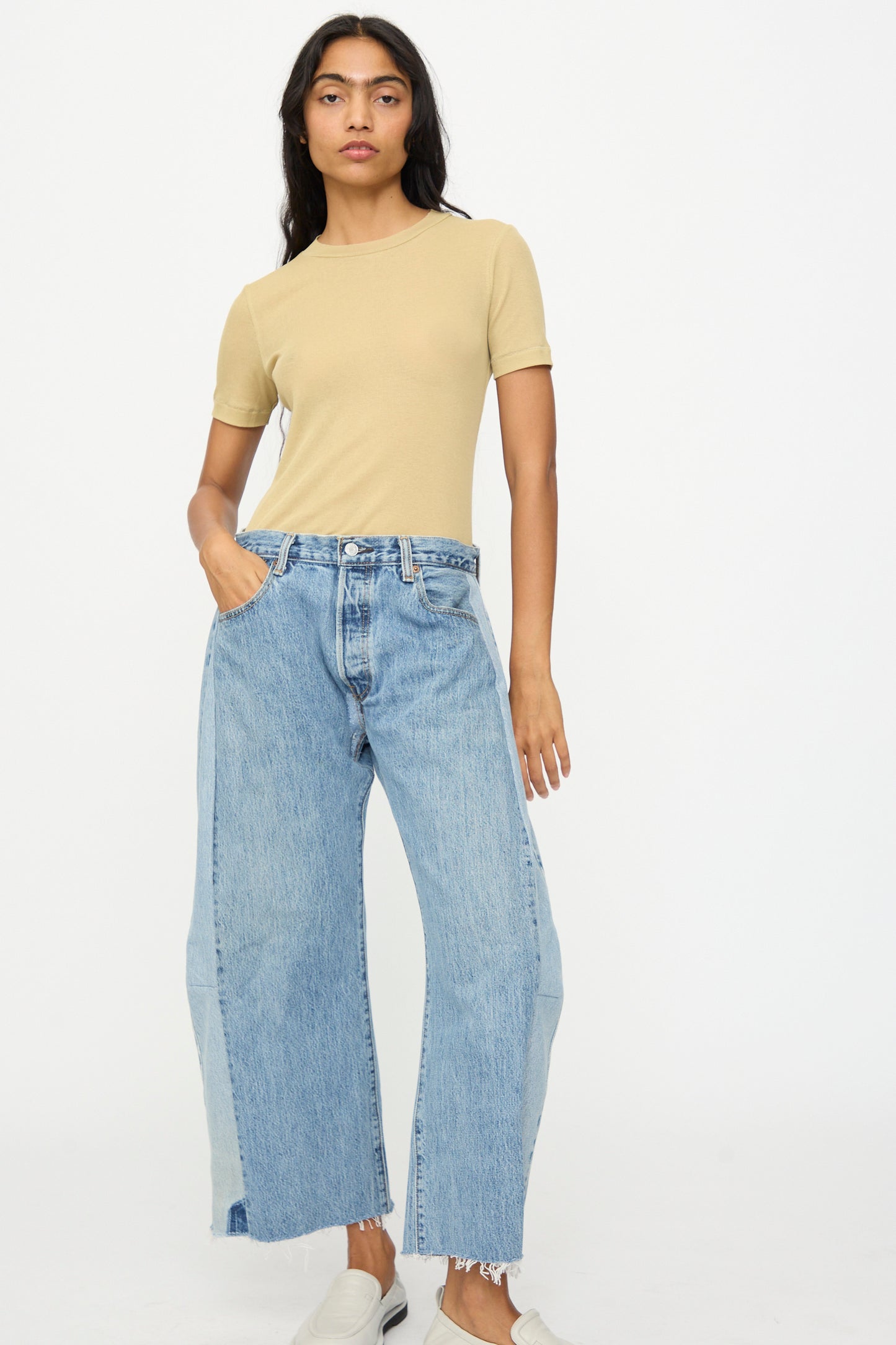 A person stands against a plain background wearing a beige short-sleeve top, B Sides Vintage Reworked Lasso Jean in Indigo, and white slip-on shoes.