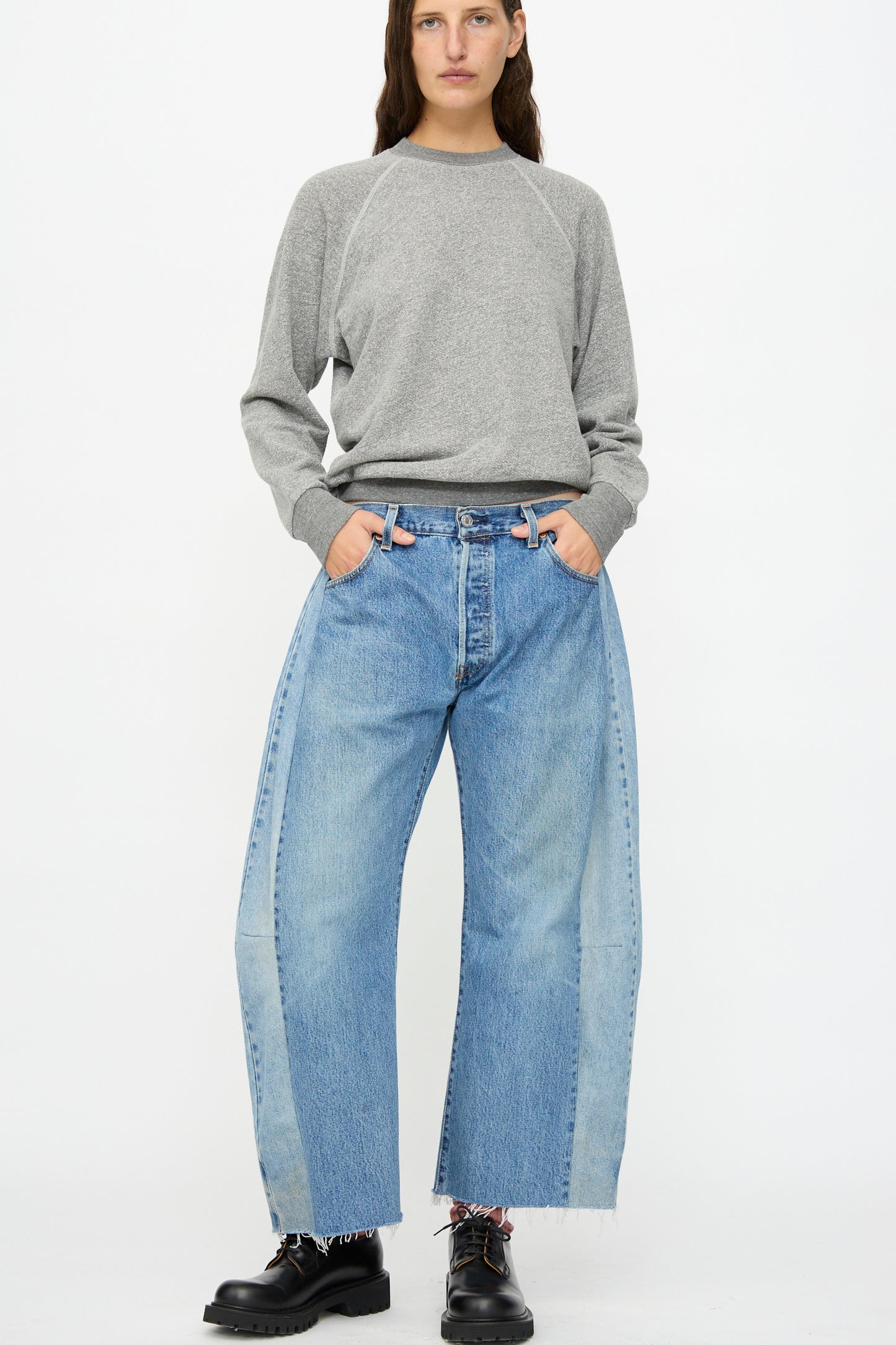 A person wearing a grey sweatshirt, B Sides Vintage Lasso Jean in Indigo, and black lace-up shoes stands against a white background with hands in pockets.