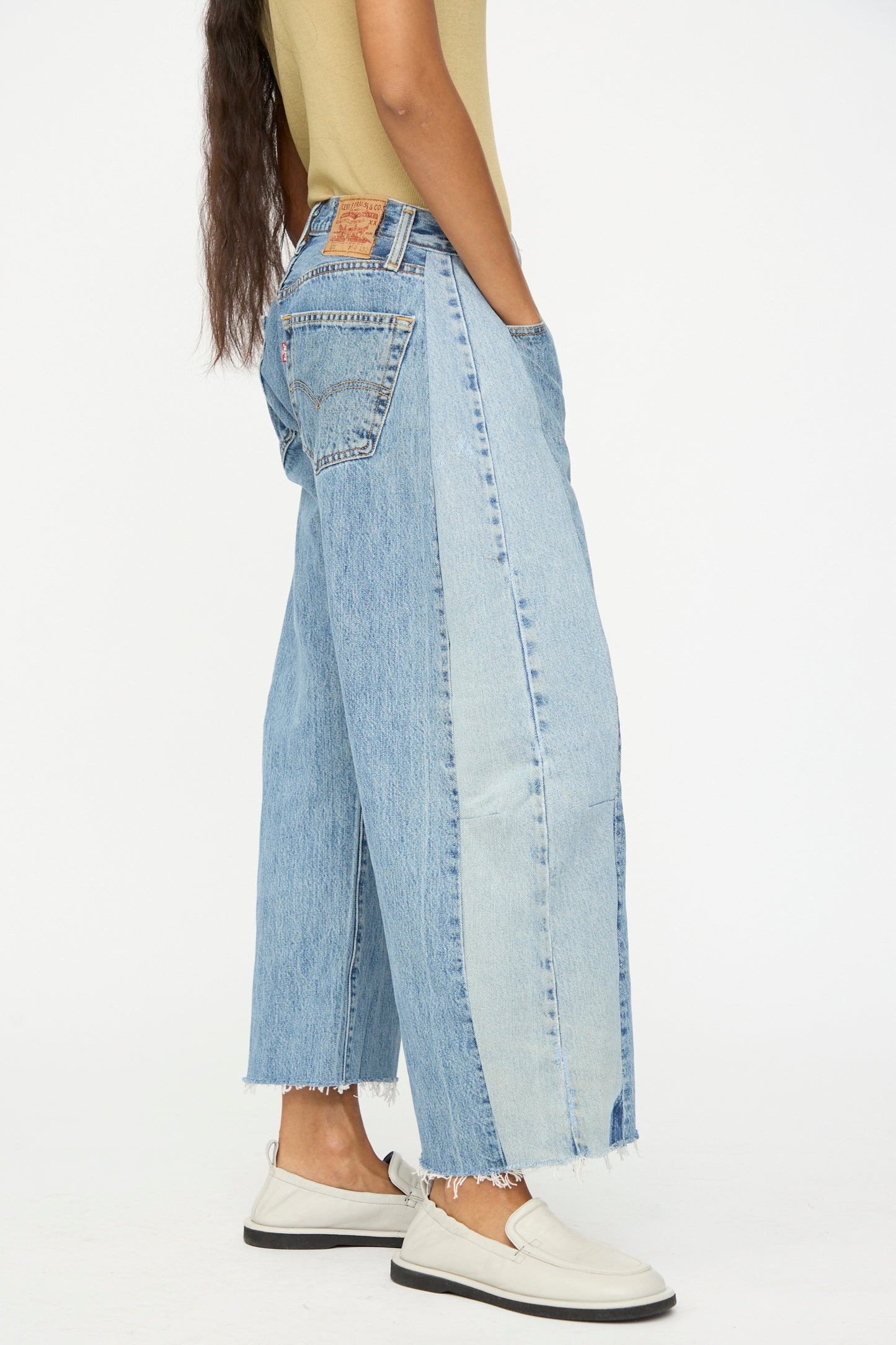 A person stands with hands in pockets, wearing B Sides Vintage Reworked Lasso Jean in Indigo, a light brown top, and white shoes against a plain background.