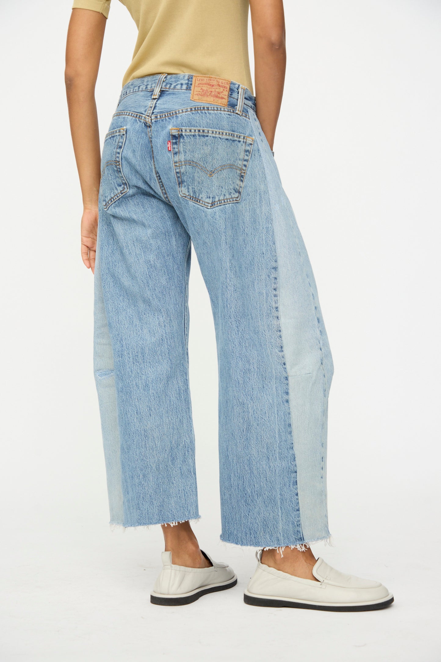 Model wears B Sides Vintage Reworked Lasso Jean in Indigo, featuring relaxed fit, wide legs, two-tone blue panels, and frayed hems, paired with white loafers against a plain white background.