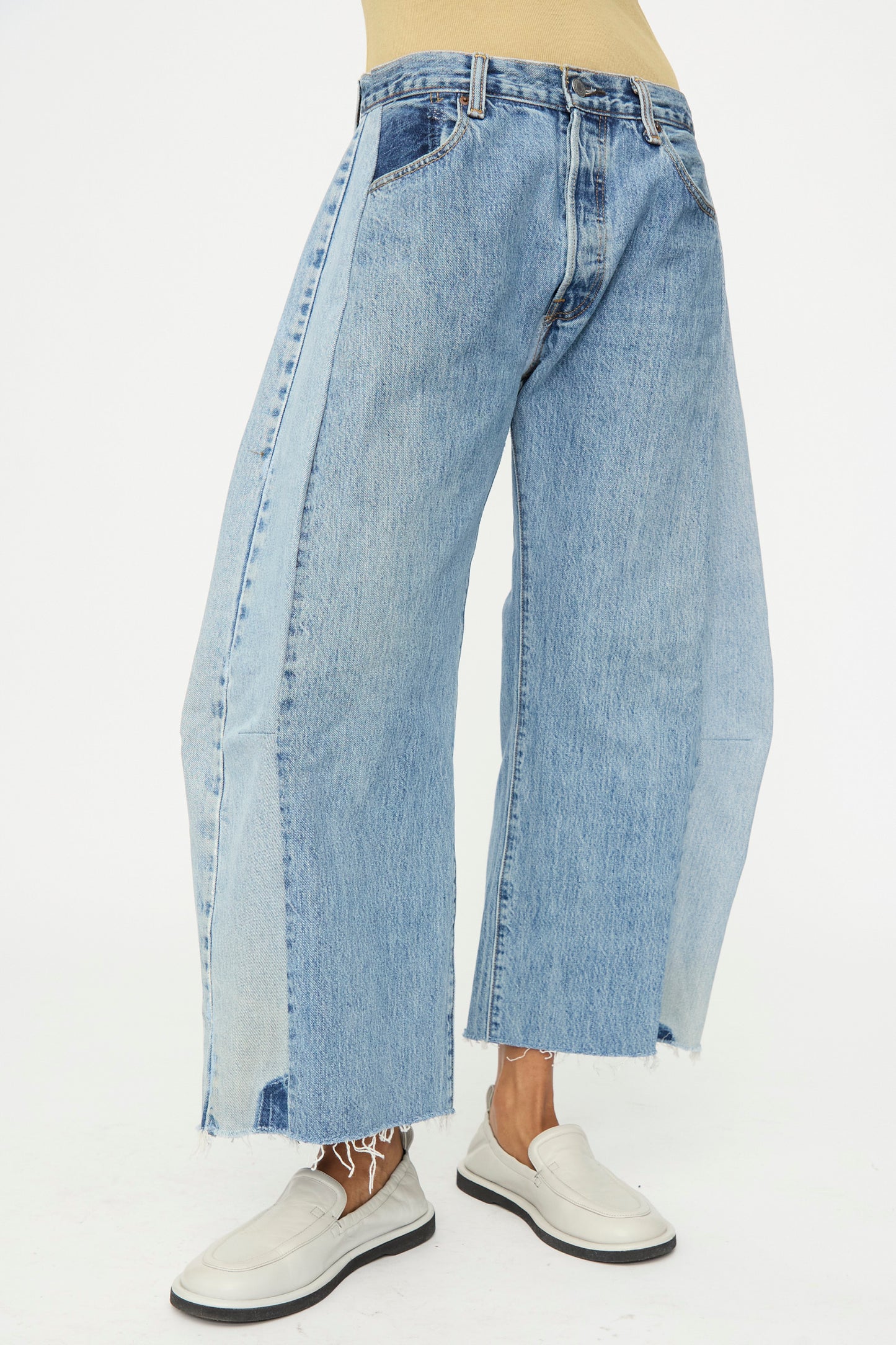 A person wears B Sides Vintage Reworked Lasso Jean in Indigo, featuring a relaxed fit, wide-leg patchwork design, and raw hems, paired with white loafers against a plain white background.