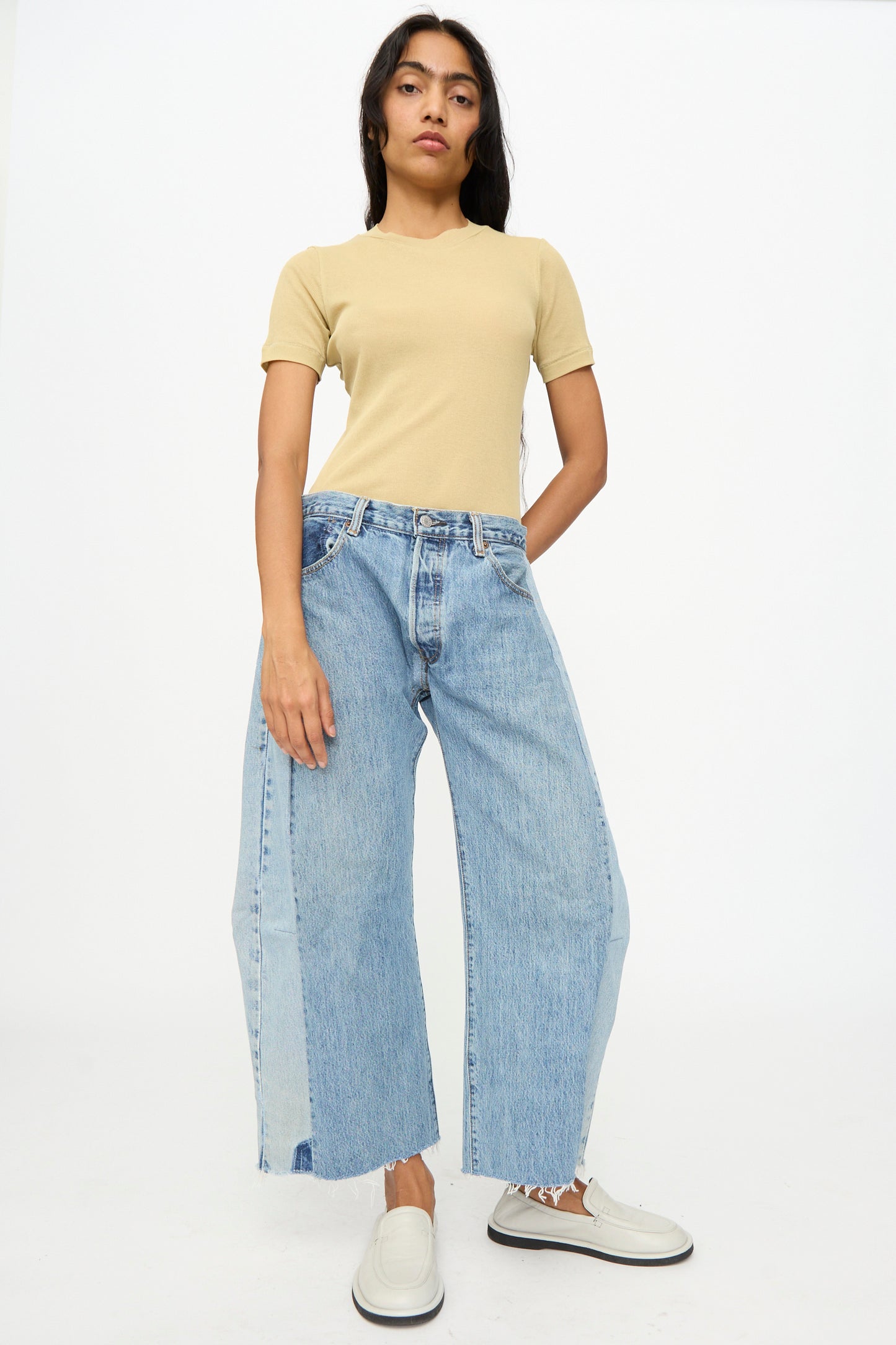 A person stands against a plain background wearing a beige t-shirt, B Sides Vintage Reworked Lasso Jean in Indigo with a relaxed fit, and white slip-on shoes.