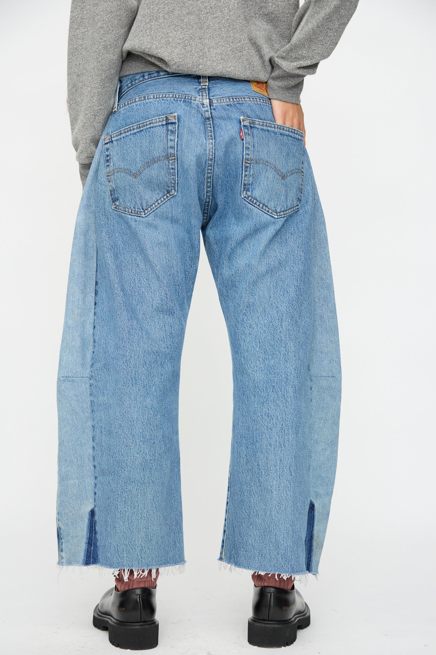 Model wears B Sides Vintage Lasso Jean in Indigo, shown from the back. Features include wide legs, two back pockets, frayed hems with slits, and a slouchy fit paired with black shoes for a relaxed look.