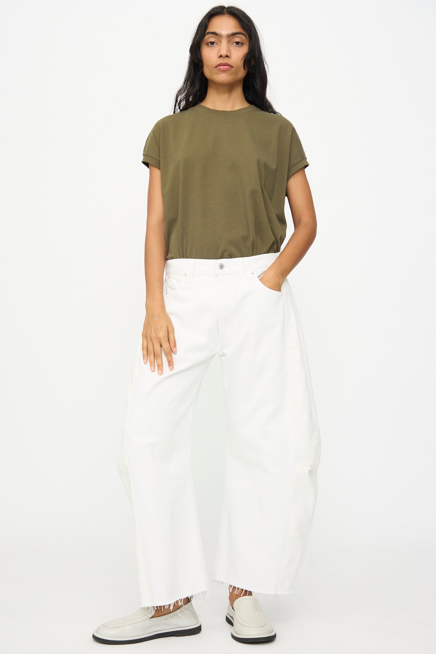 Person standing against a plain background, wearing an olive green t-shirt, B Sides Vintage Reworked Lasso Jean in White, and light slip-on shoes, with one hand in their pocket.