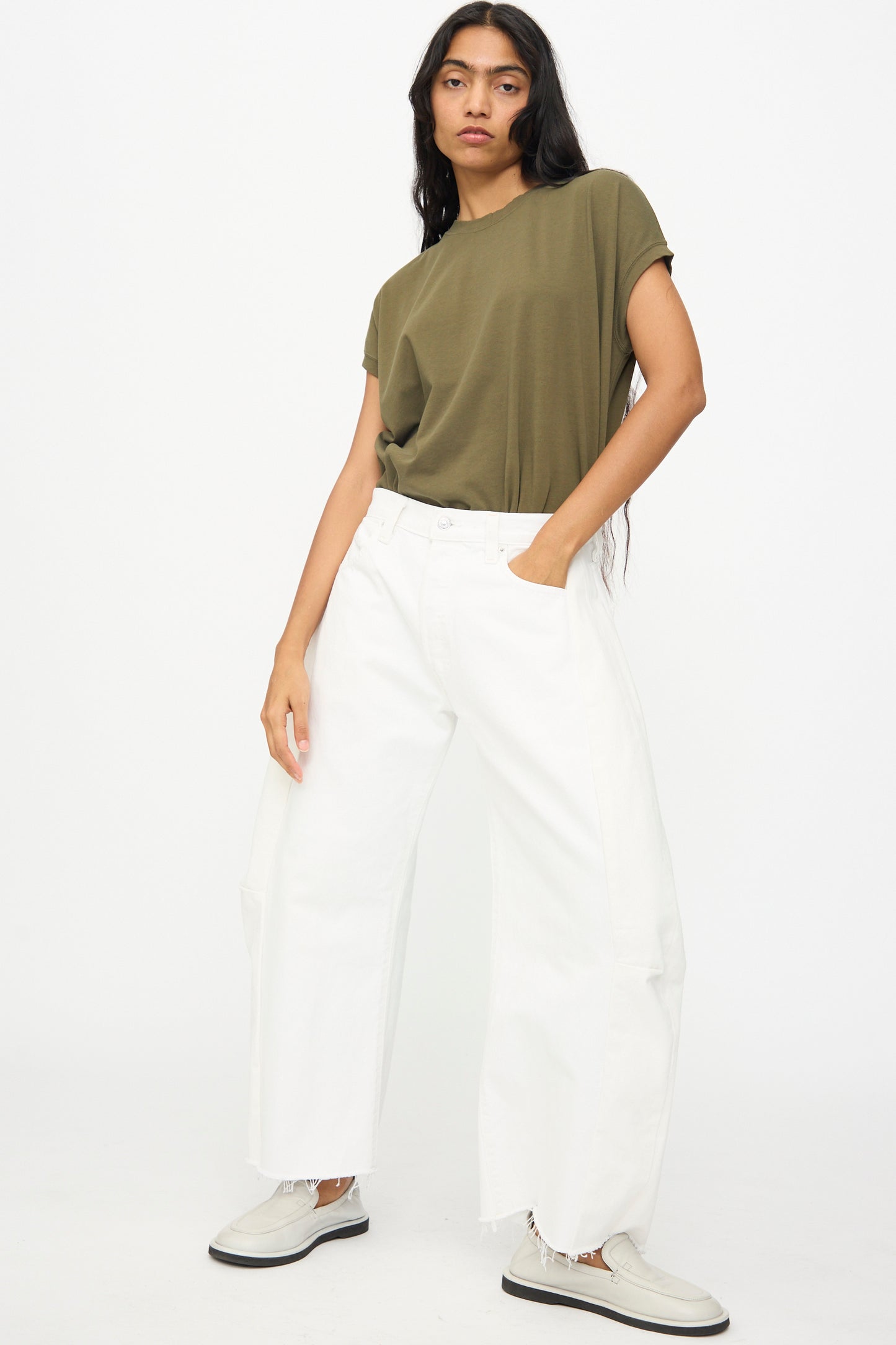 A person stands against a plain background wearing an olive green t-shirt, B Sides Vintage Reworked Lasso Jean in White, and white slip-on shoes, with one hand in their pocket.