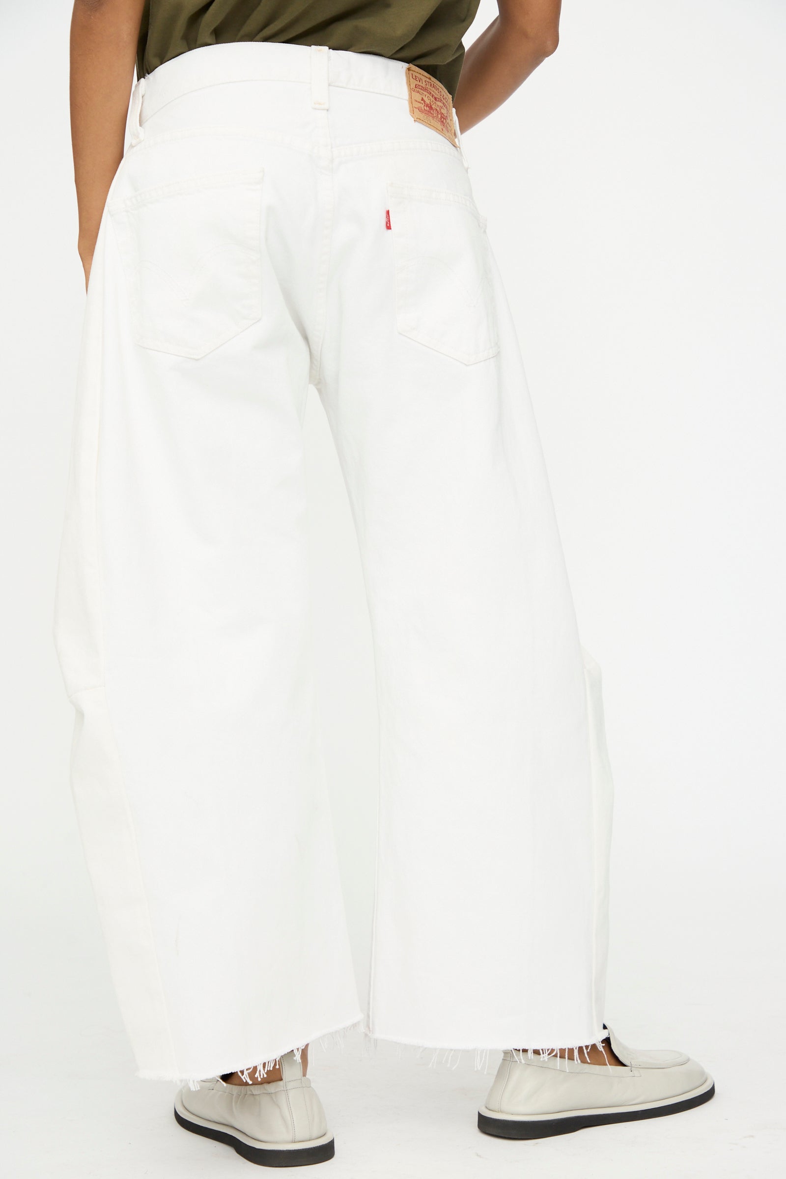 Model wears B Sides Vintage Reworked Lasso Jean in White with frayed hems, white shoes, and a dark green top against a plain white background.