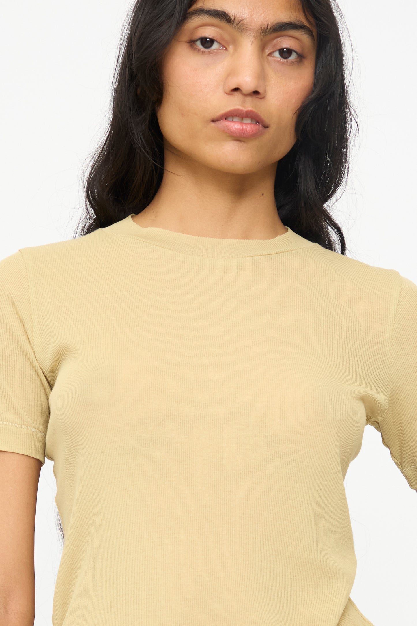 A person with long dark hair wears the B Sides Vivien Rib Tee in Honey, a slim fit, short-sleeved beige top, and looks toward the camera against a plain white background.
