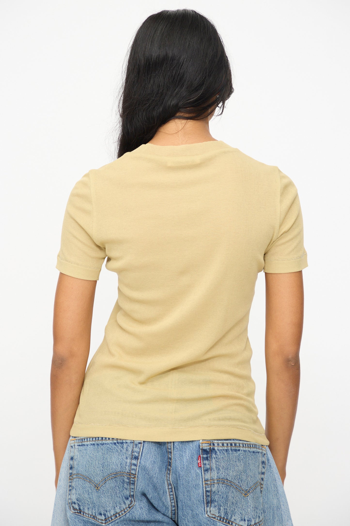 A person with long dark hair, seen from the back against a plain white background, wears the B Sides Vivien Rib Tee in Honey and blue denim jeans.