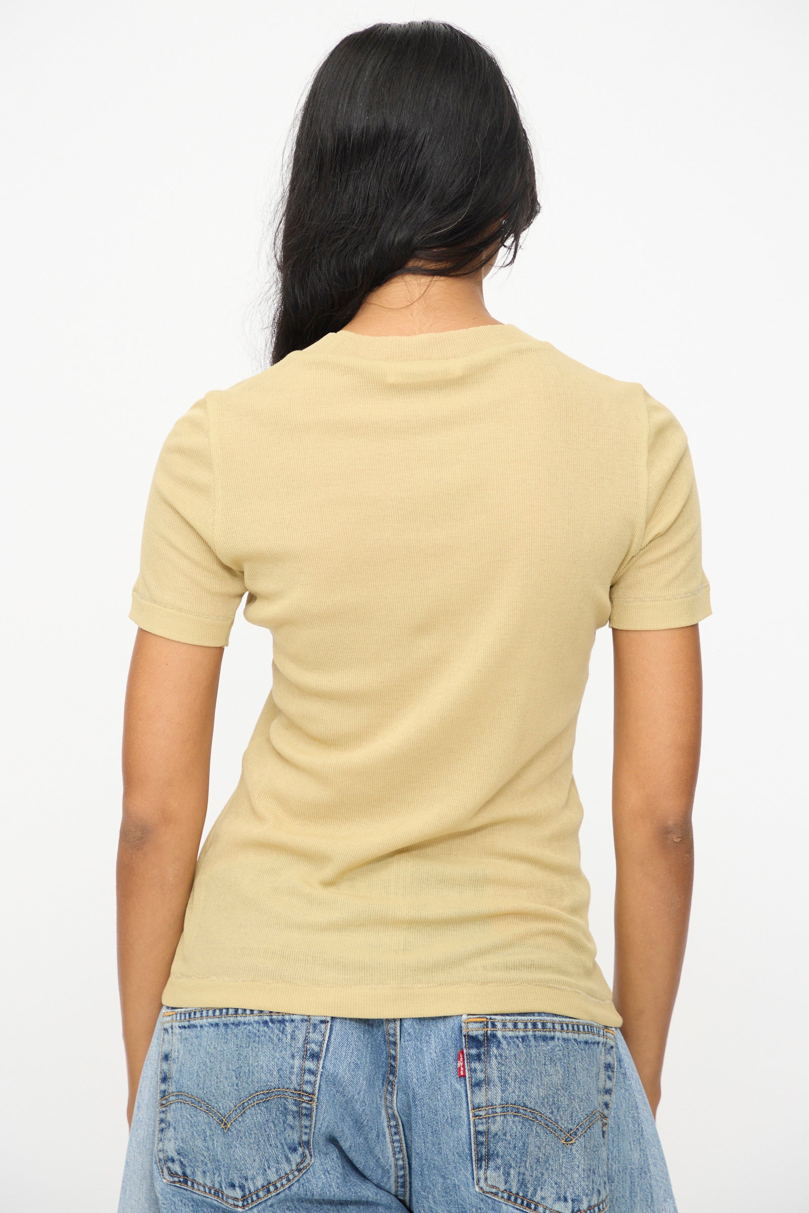 A person with long dark hair, seen from the back against a plain white background, wears the B Sides Vivien Rib Tee in Honey and blue denim jeans.