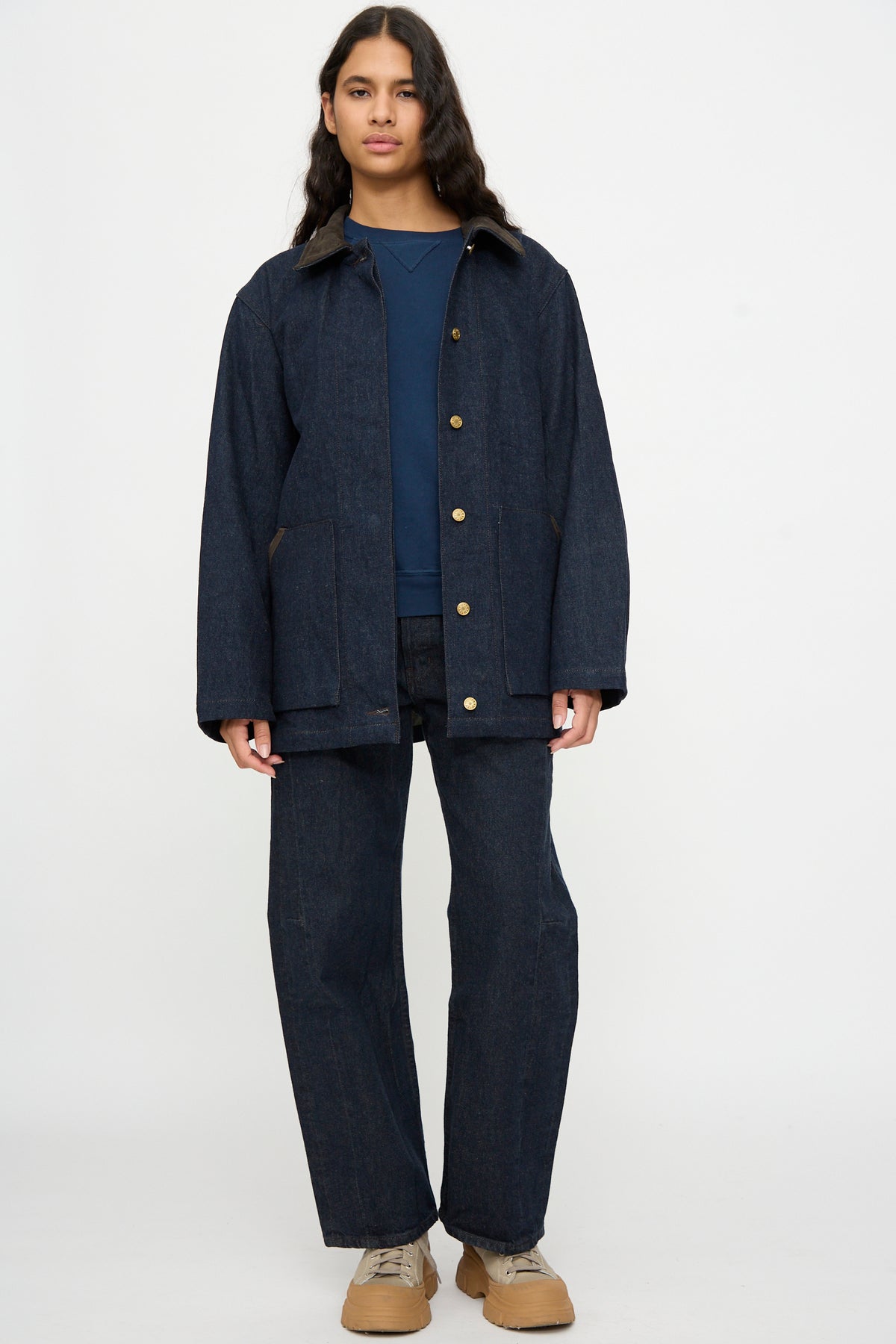 A person stands against a plain background wearing the B Sides Workwear Jacket in Indigo Rinse and Espresso Suede, matching denim pants, a blue shirt, and beige sneakers.