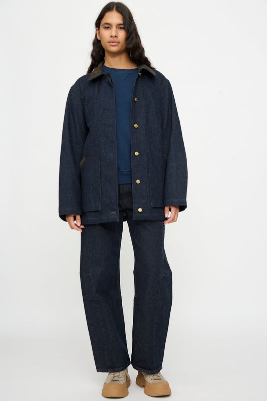 A person stands against a plain background wearing the B Sides Workwear Jacket in Indigo Rinse and Espresso Suede, matching denim pants, a blue shirt, and beige sneakers.