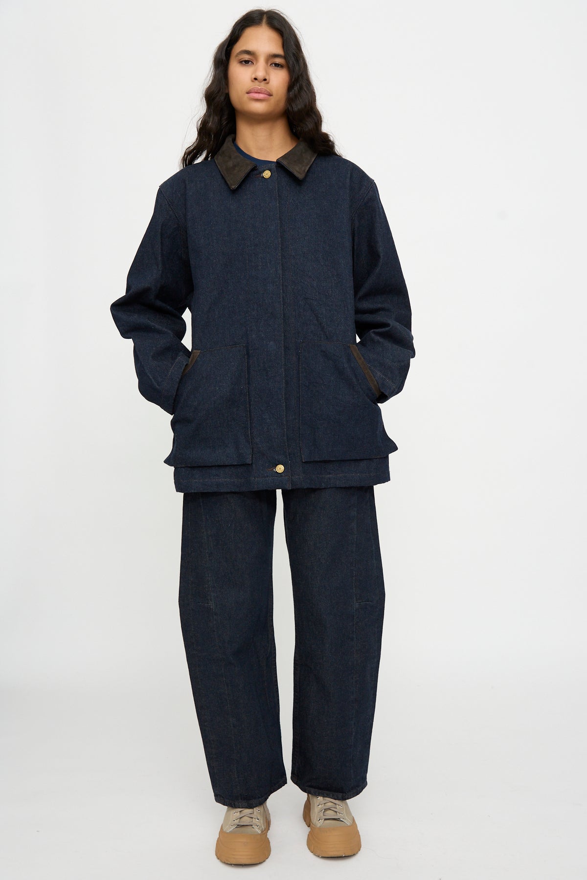 Person stands against a plain background, wearing the B Sides Workwear Jacket in Indigo Rinse and Espresso Suede with matching pants, tan shoes, hands in pockets, looking ahead.