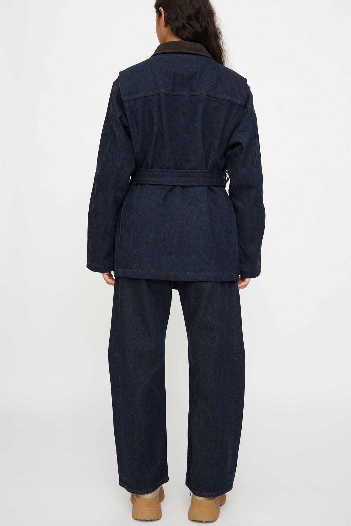 Person facing away, wearing the B Sides Workwear Jacket in Indigo Rinse and Espresso Suede, wide-leg pants, and tan shoes against a white background.