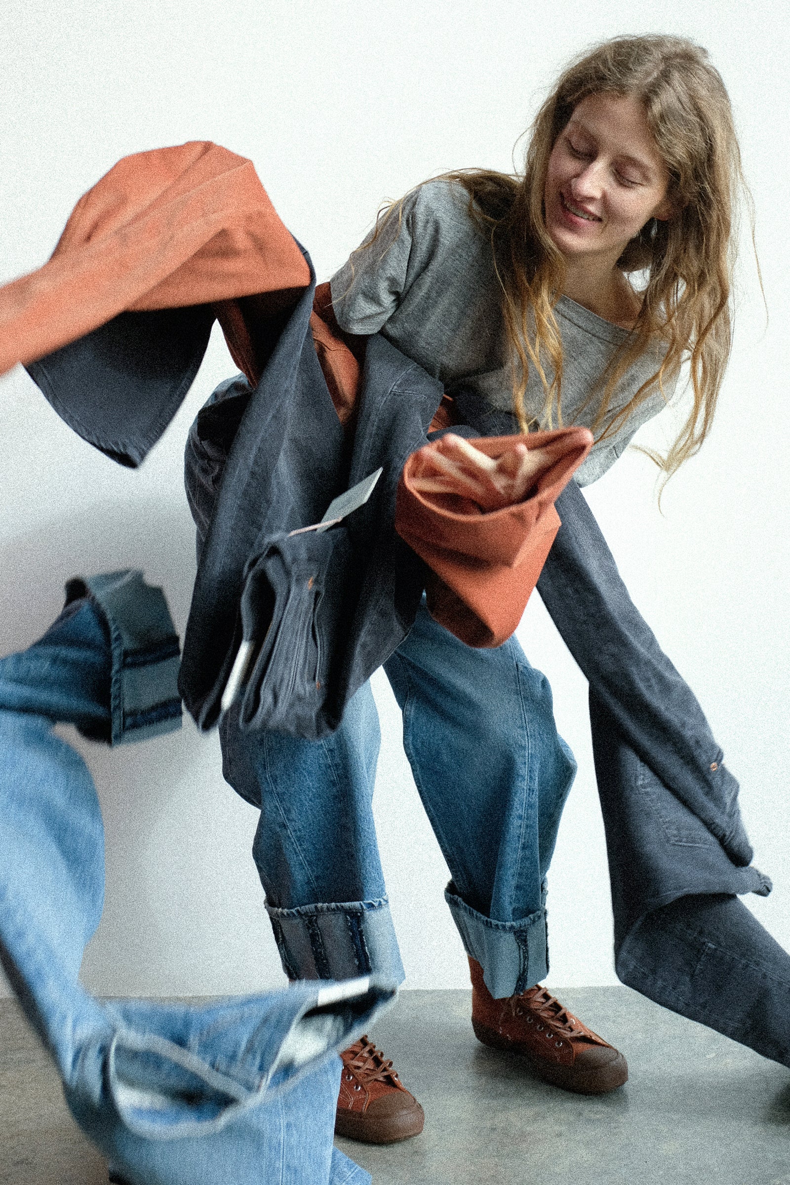A woman trying to hold up B Sides jeans against a white wall.