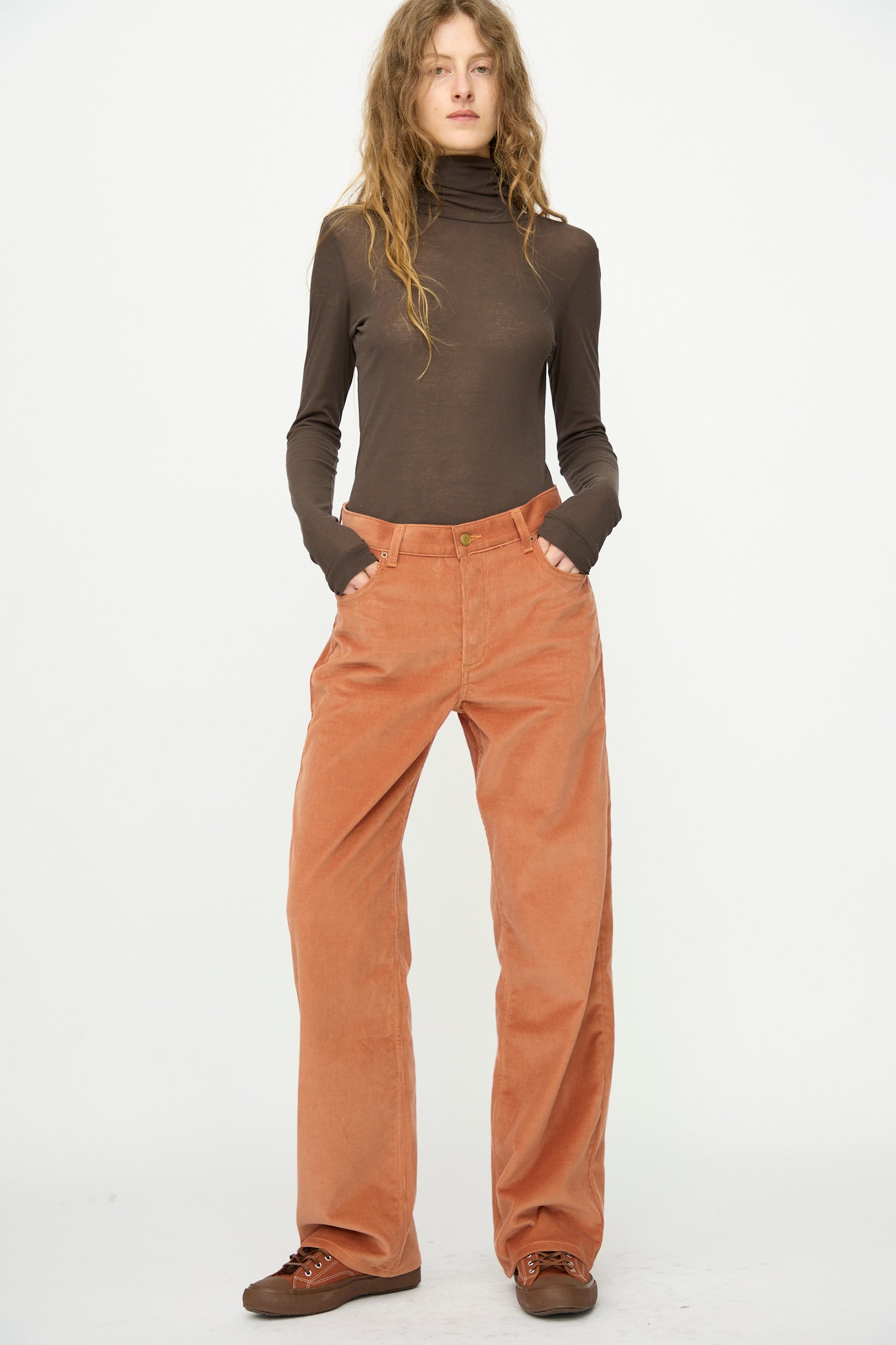 A person stands against a plain background wearing B Sides Bec Jean in Peach Tile, a brown turtleneck, and brown shoes with hands in their pockets.
