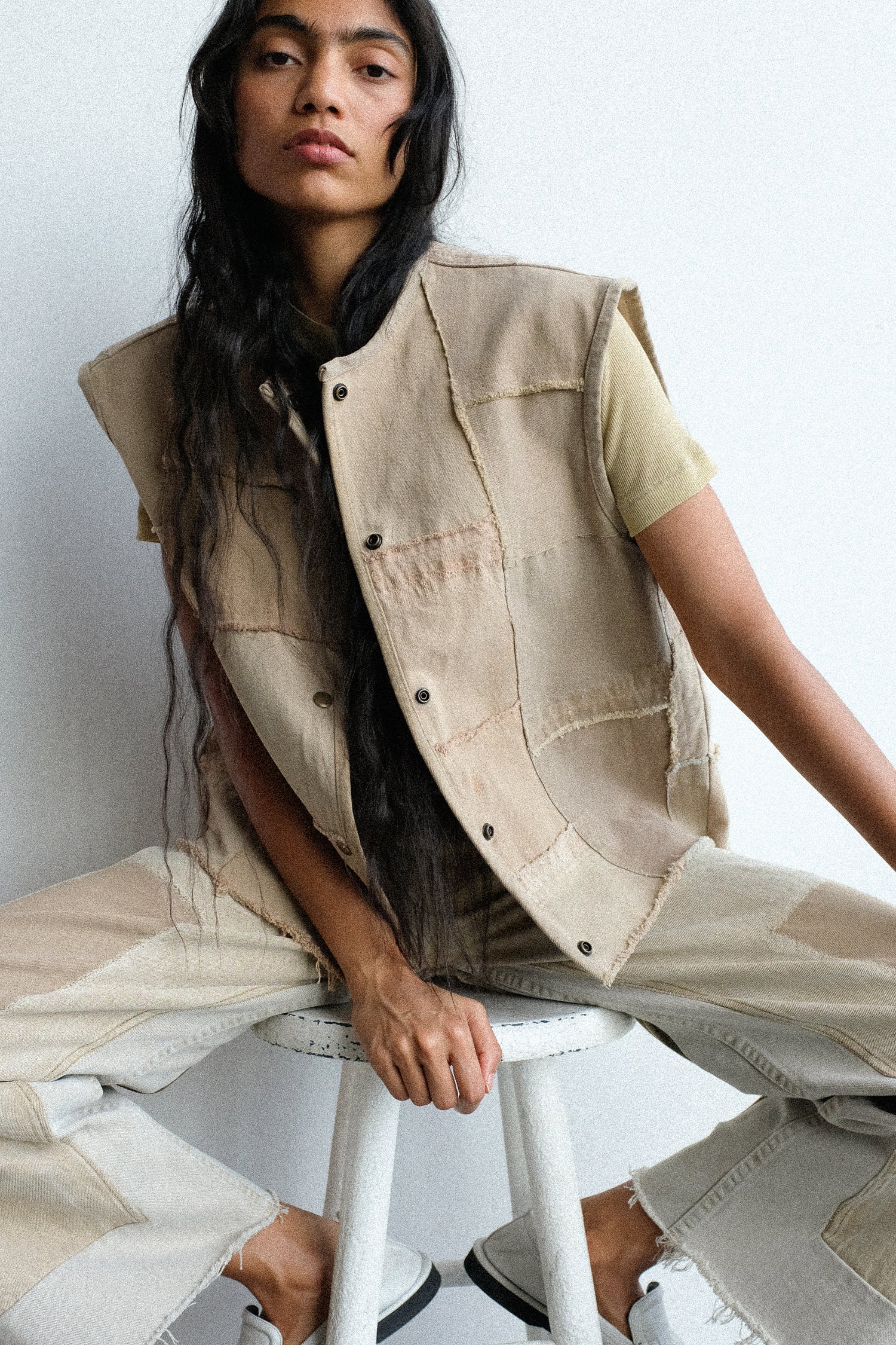 Person wearing a beige vest and pants sitting on a white stool against a plain background