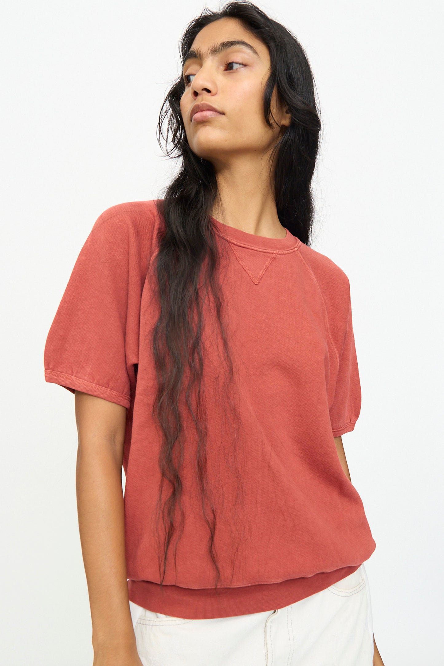 A person with long wavy hair stands against a plain white background wearing the B Sides Short Sleeve Sweatshirt in Faded Tomato Overdye, a relaxed fit top crafted from soft French terry.