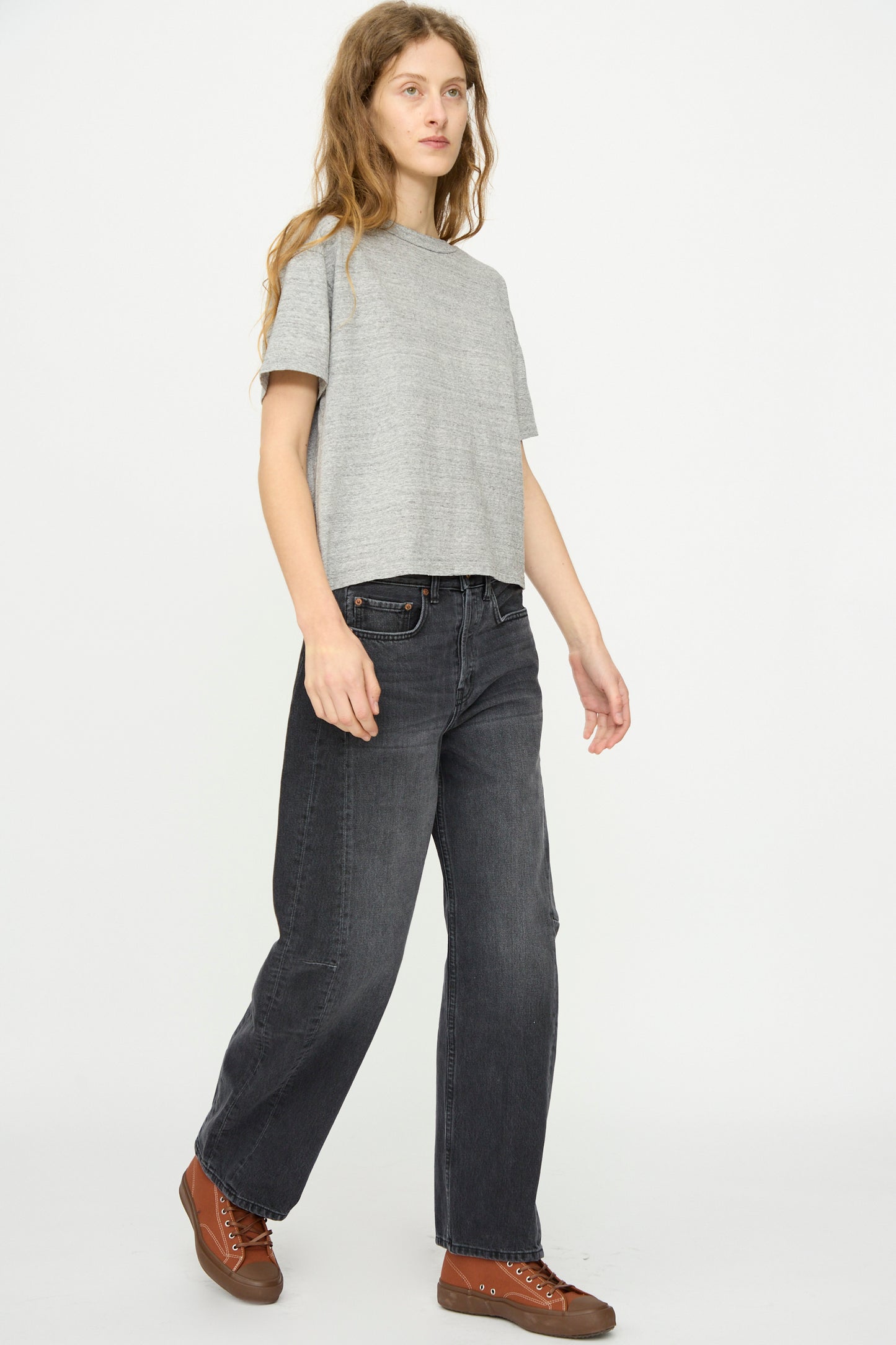 A person with long hair wears a gray short-sleeve T-shirt, B Sides Slim Lasso Jean in Stil Black, and brown lace-up shoes while walking against a plain white background.