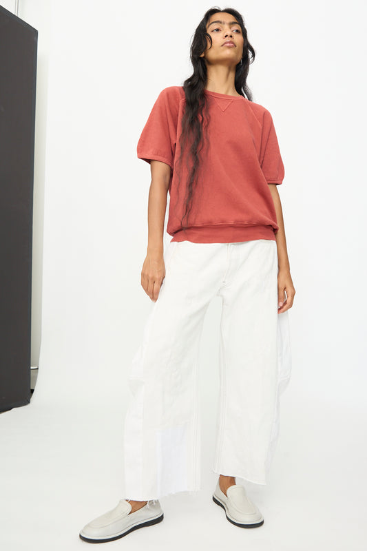 A person stands against a plain background, wearing a red short-sleeve sweatshirt, light gray slip-on shoes, and B Sides' Vintage Reworked Lasso Jean in White.