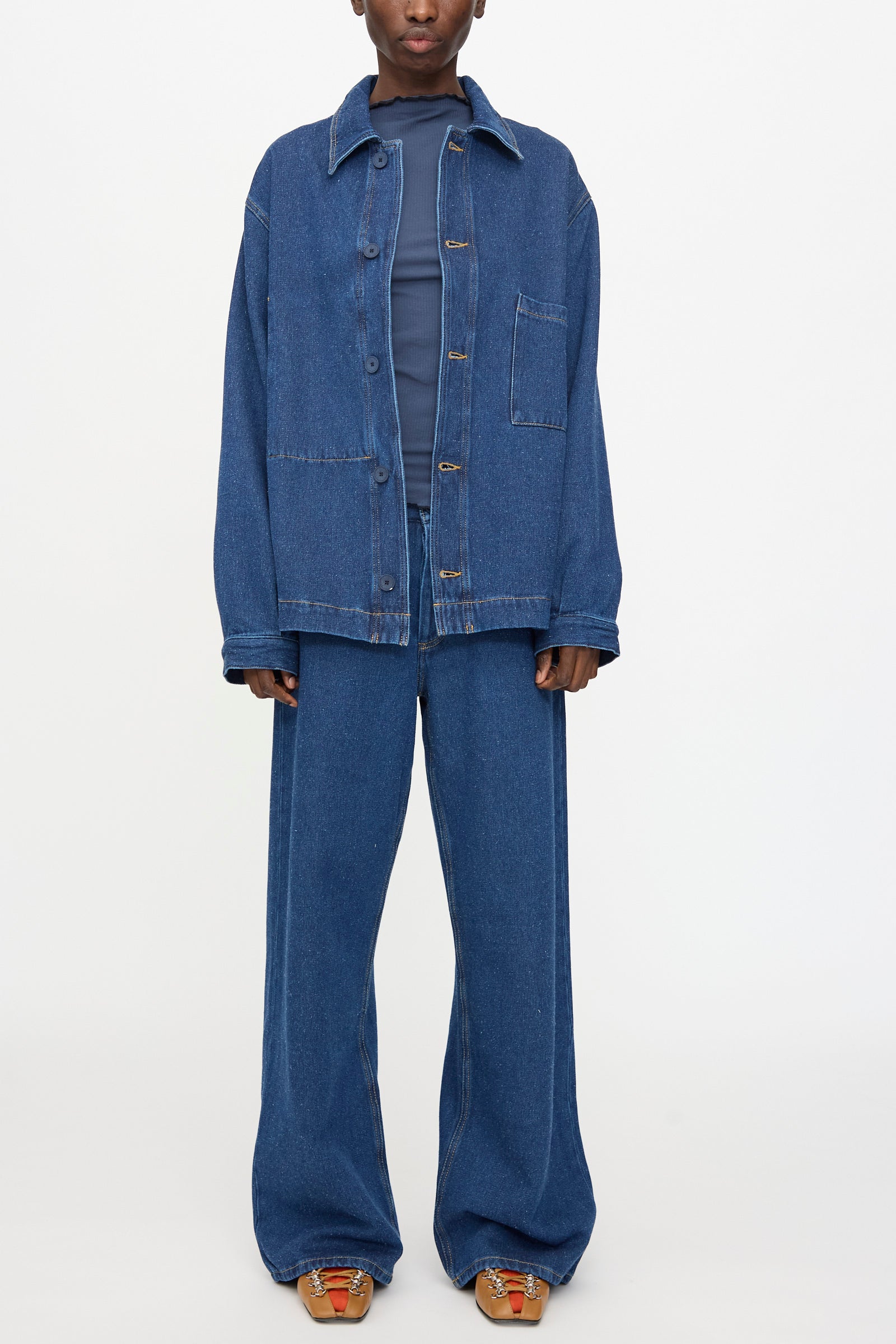 Wearing the Baserange Altru Jacket in Indigo Blue with wide-leg jeans over a dark shirt, a person stands against a plain background.