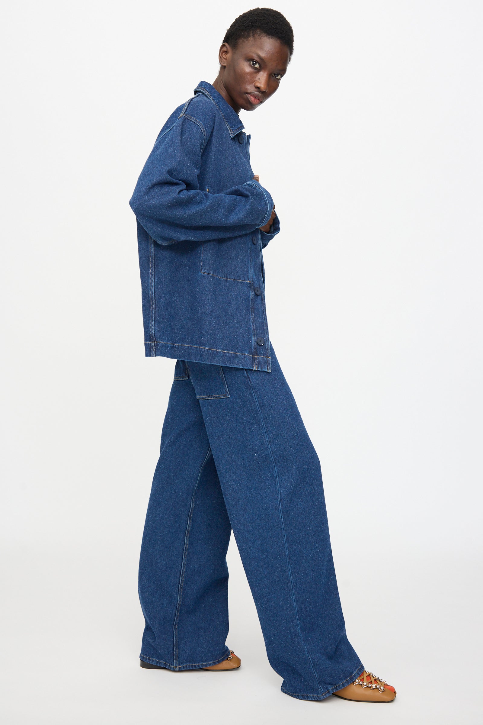 Model wears the Baserange Altru Jacket in Indigo Blue and relaxed fit wide-leg denim jeans, standing sideways against a plain white background.