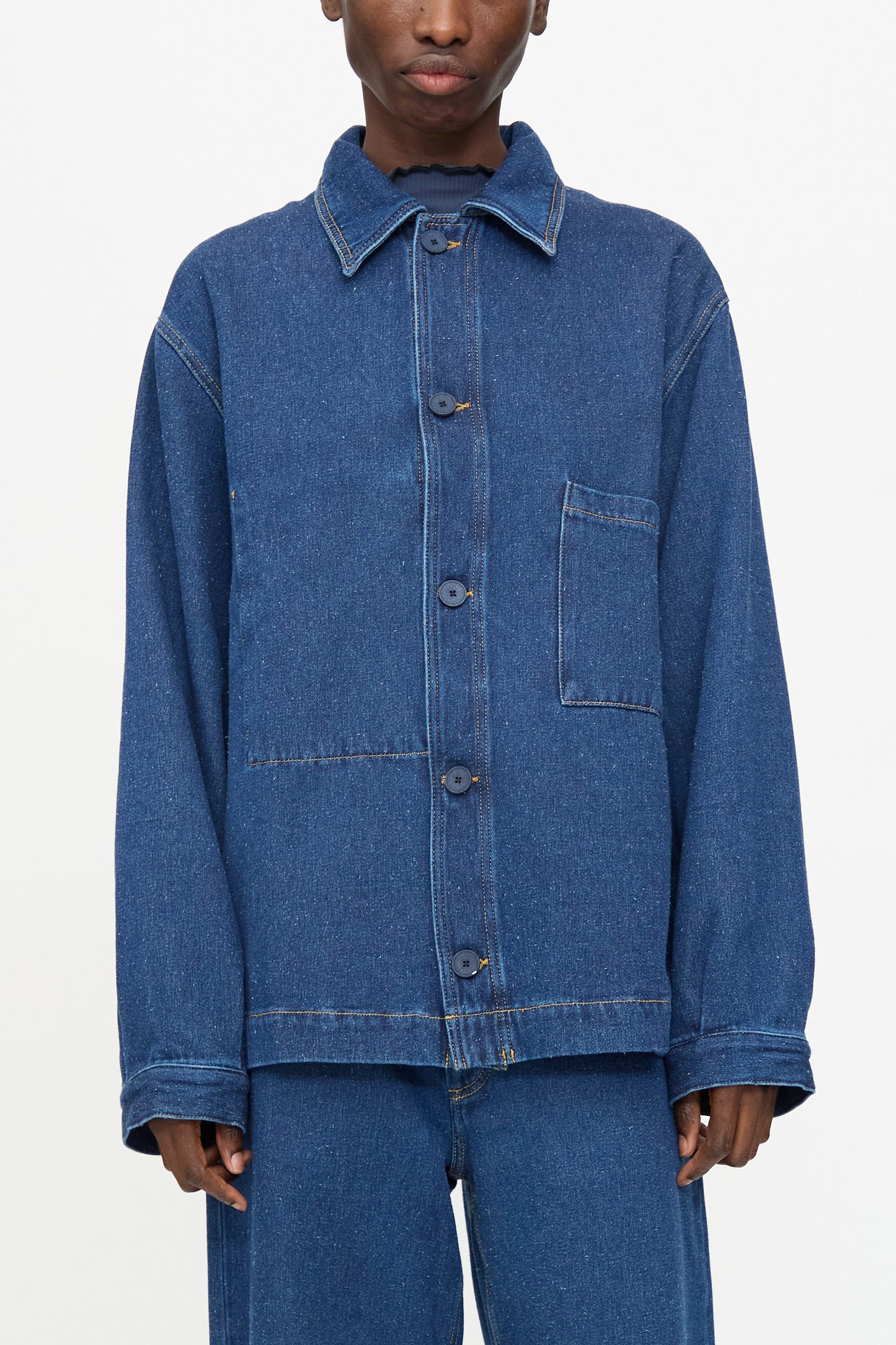 Model wearing the Baserange Altru Jacket in Indigo Blue, featuring visible seams, front buttons, a single chest pocket, and paired with matching denim pants against a plain background.
