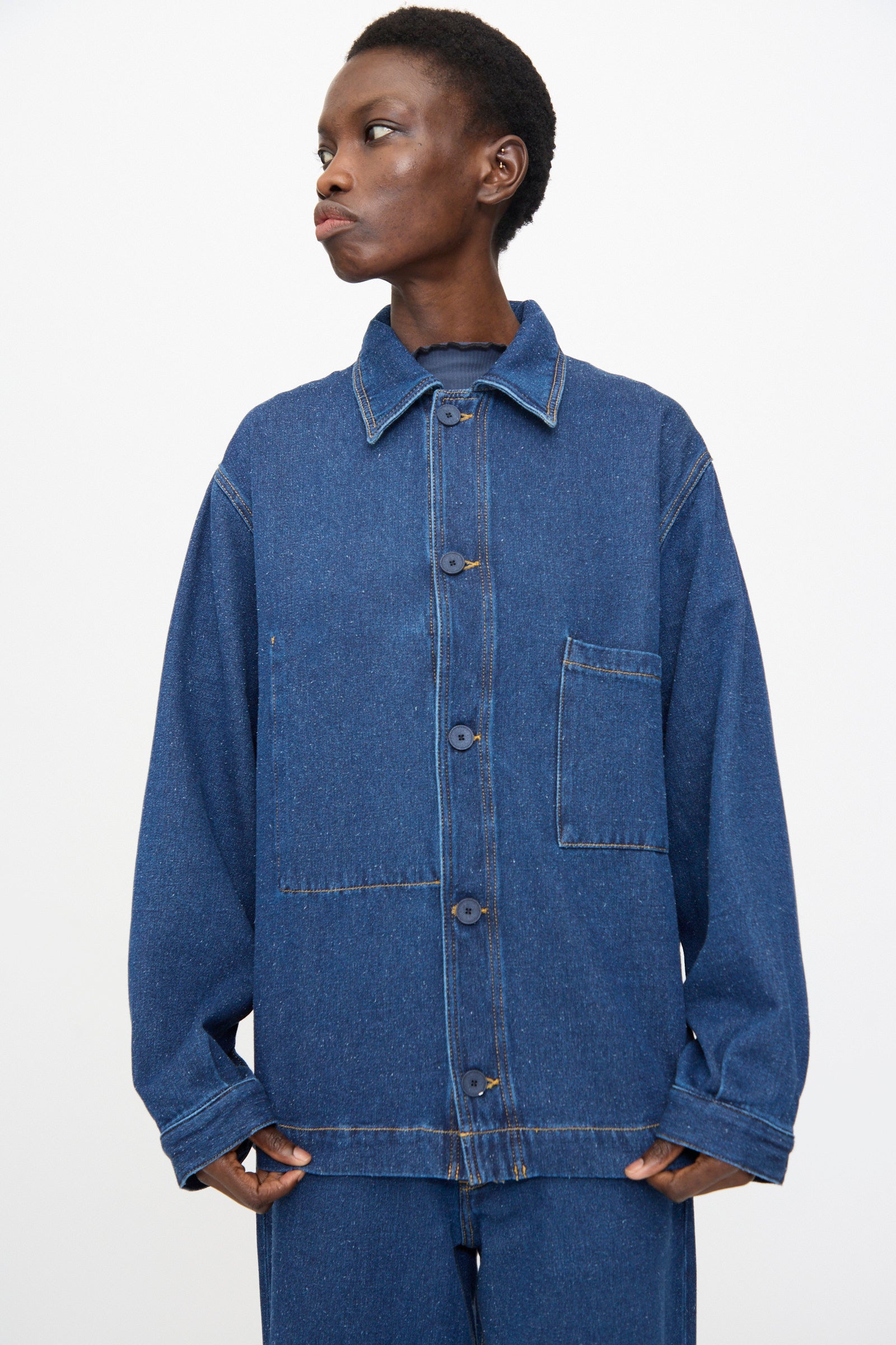 A person wears the Baserange Altru Jacket in Indigo Blue—a relaxed fit, button-up jacket with a chest pocket—paired with matching denim pants, standing against a plain white background.