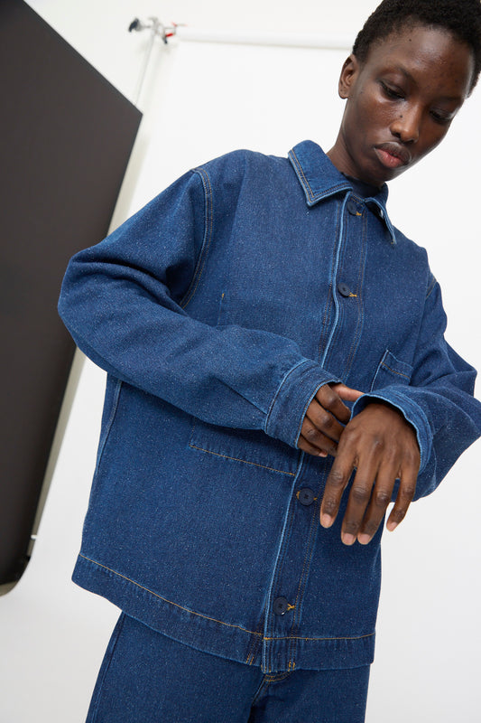 A person wears the Baserange Altru Jacket in Indigo Blue, paired with jeans, standing indoors against a white background with a black panel on the left.