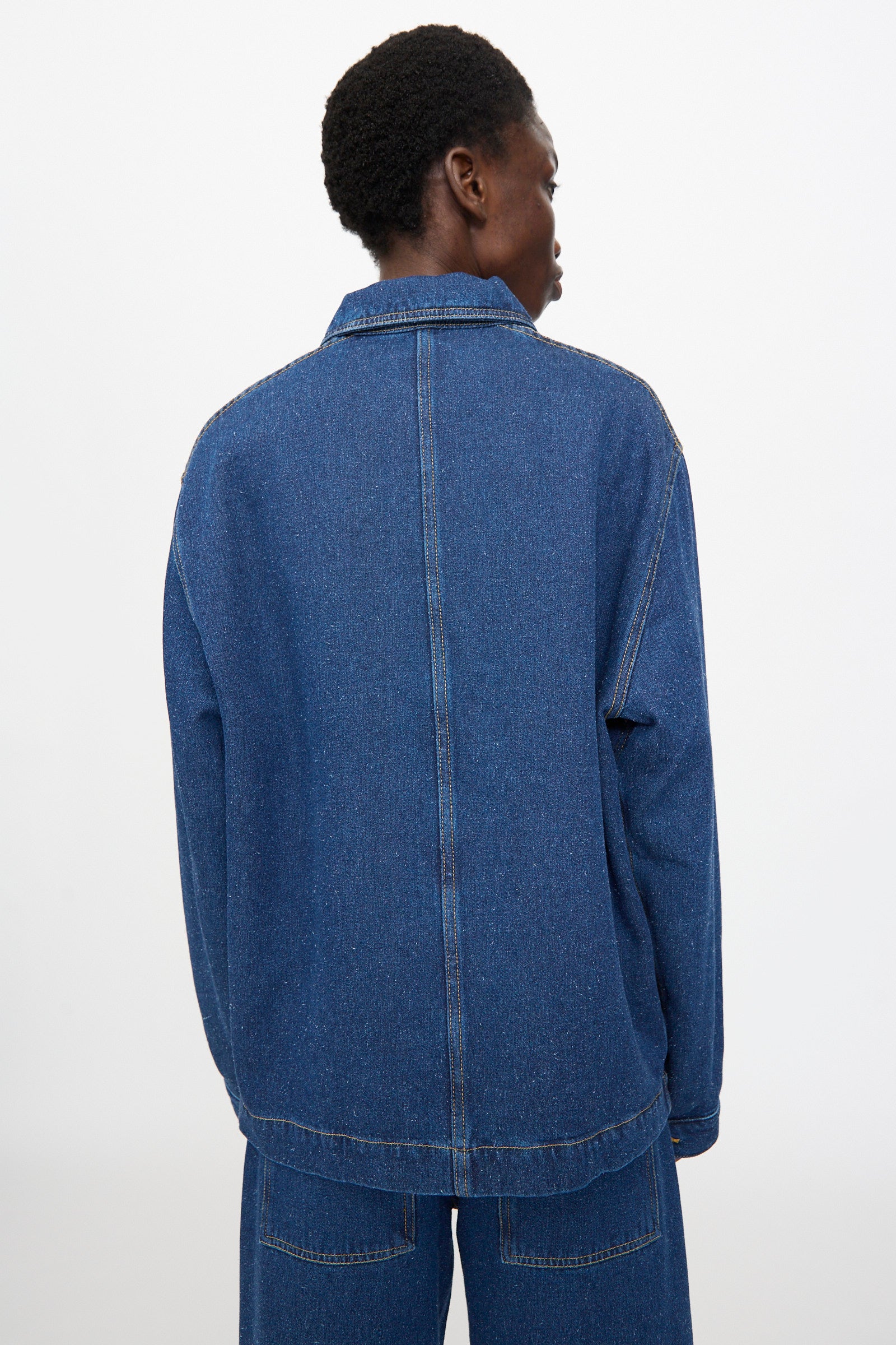 A person is shown from the back against a plain white background wearing the Baserange Altru Jacket in Indigo Blue, styled with matching denim pants.
