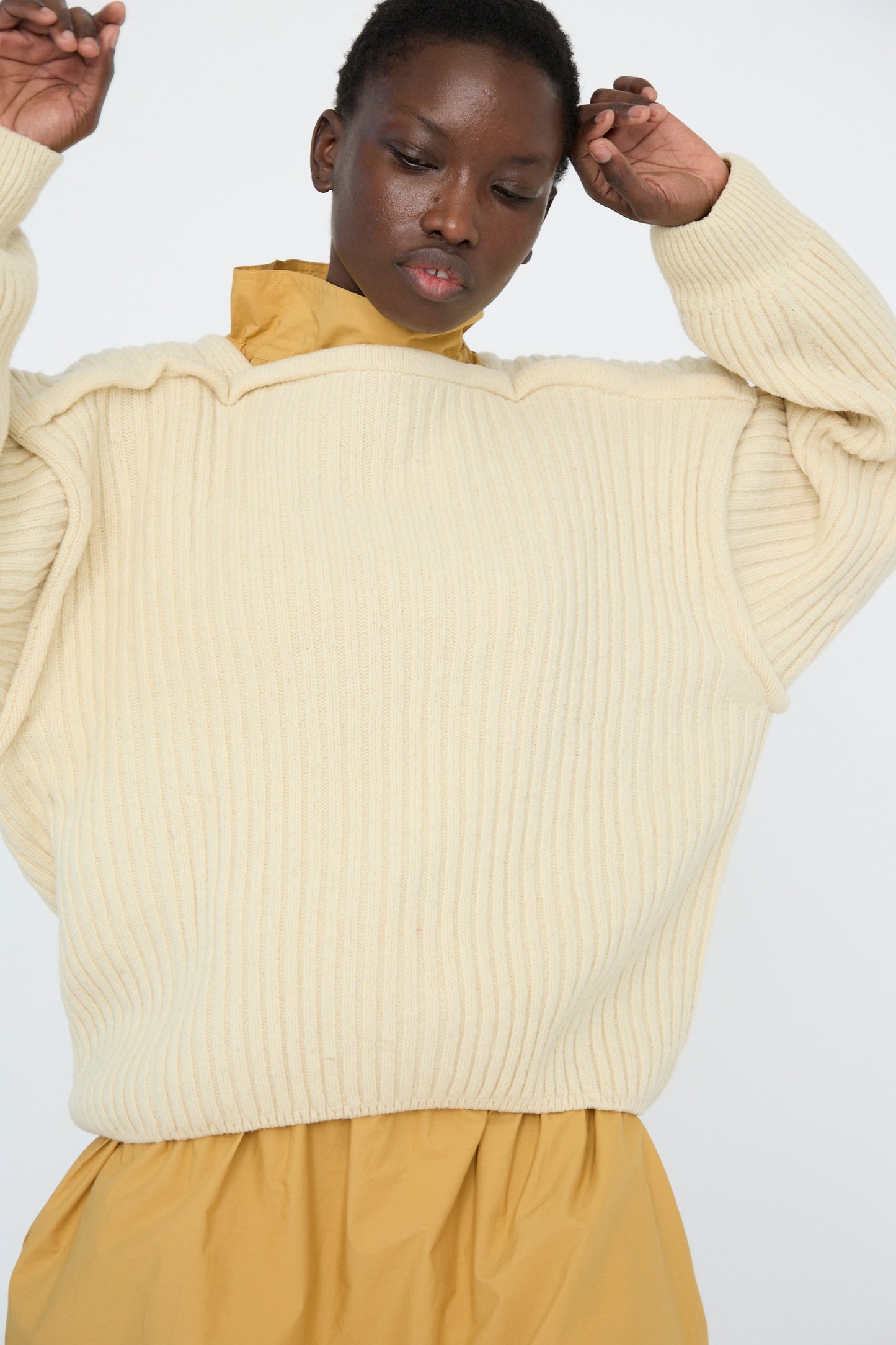 A person wearing a Baserange Arles Merino Wool Construct Sweater in Ecru over a mustard shirt stands against a plain background, arms raised.