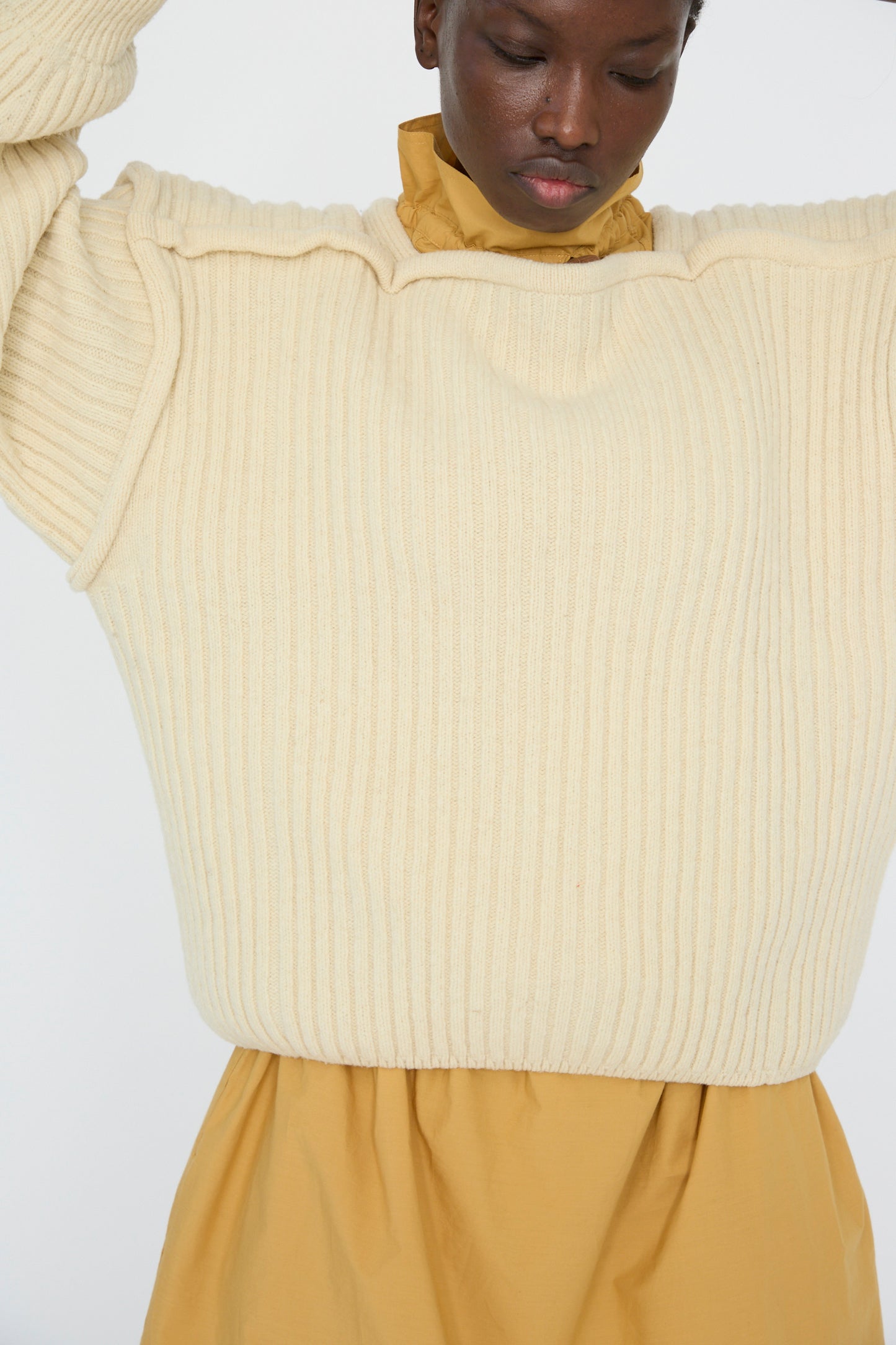 A person in a Baserange Arles Merino Wool Construct Sweater in Ecru raises their arms, with the cream sweater's vertical ribbing layered over a mustard yellow dress.