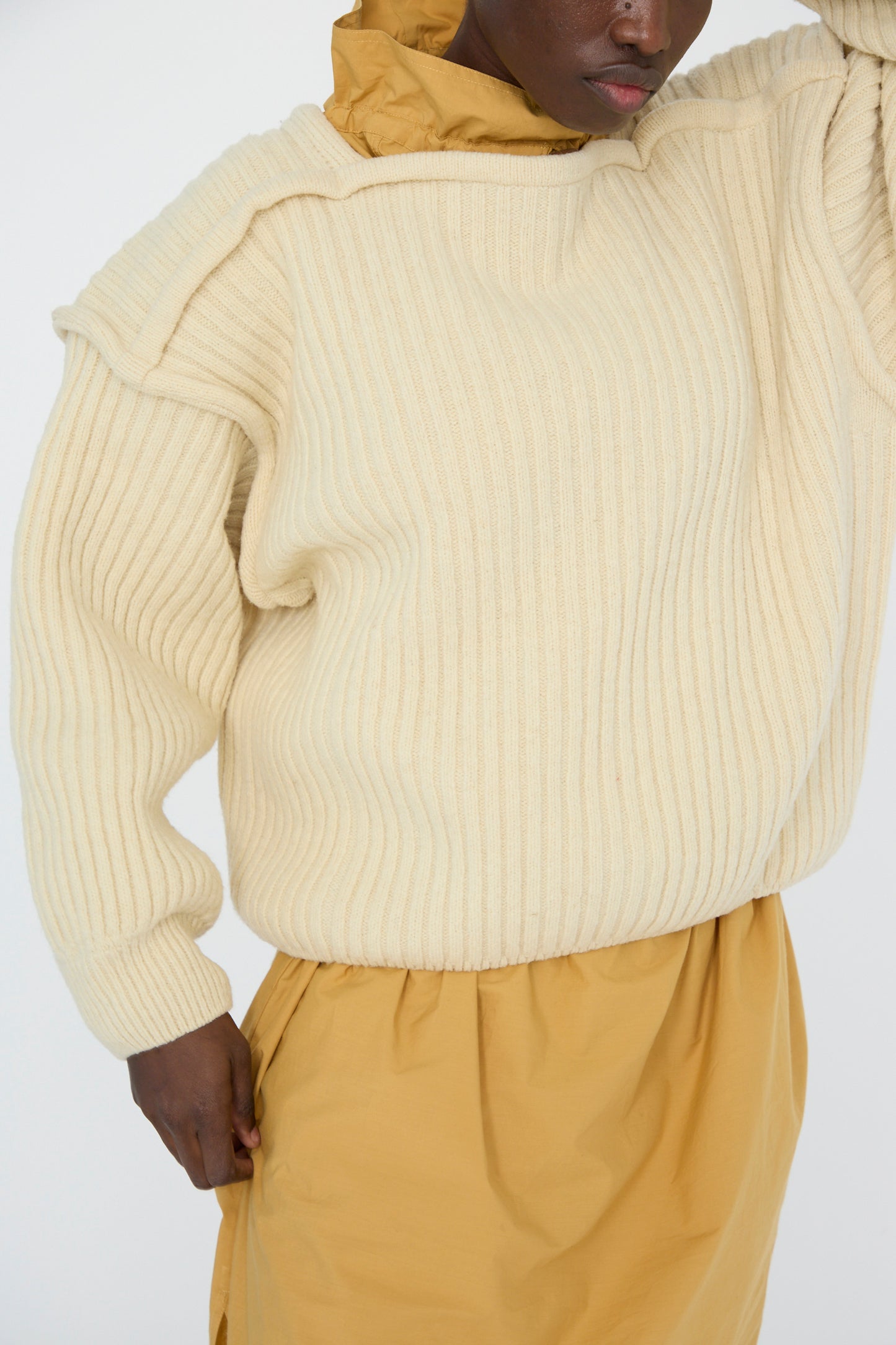 A person wears an oversized Baserange Arles Merino Wool Construct Sweater in ecru, paired with a mustard yellow dress. The sweater's cream vertical ribbing adds texture against the plain background.