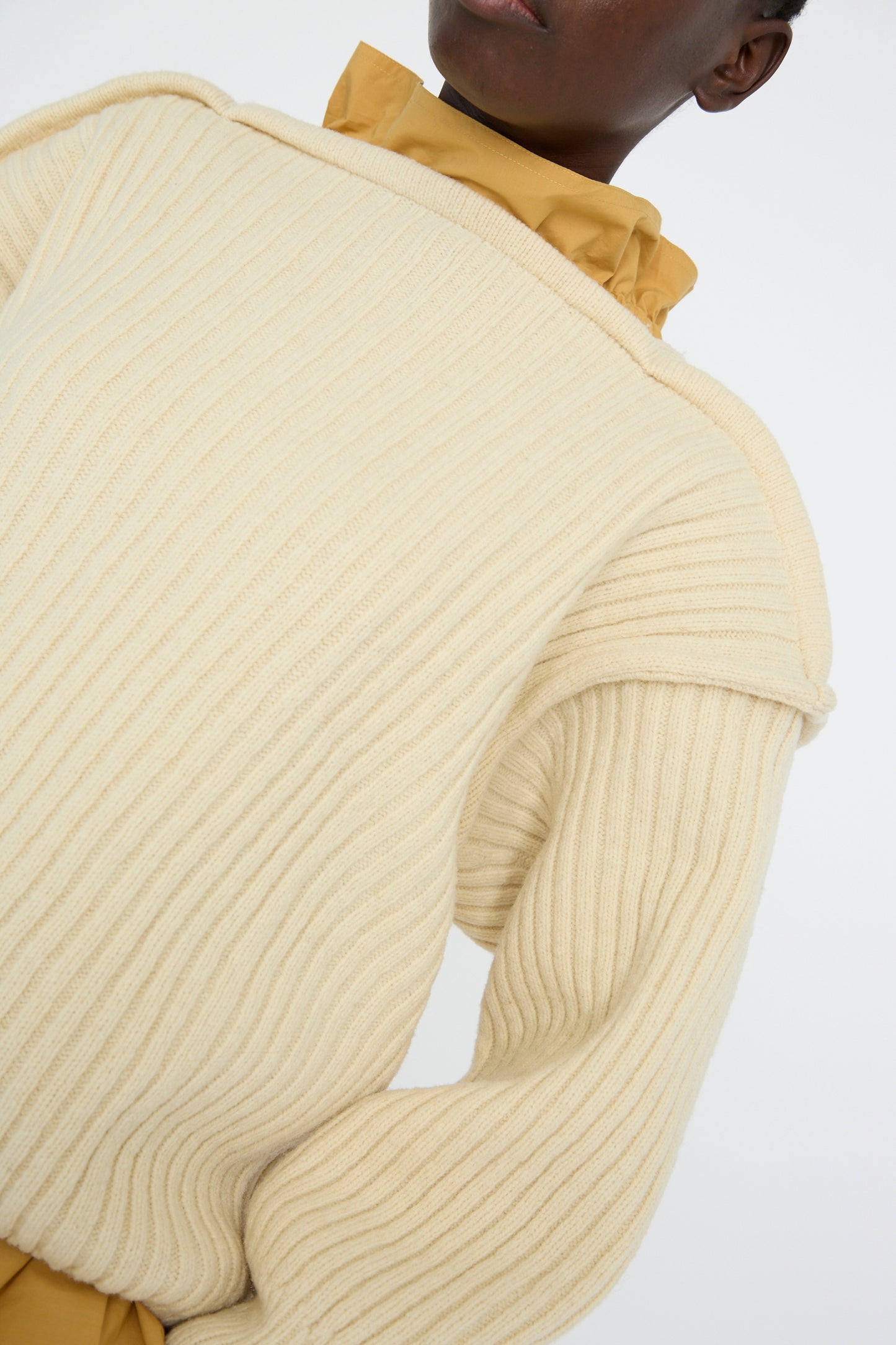 Close-up of someone in a Baserange Arles Merino Wool Construct Sweater in Ecru, highlighting its vertical ribbing texture over a mustard turtleneck against a white backdrop. 