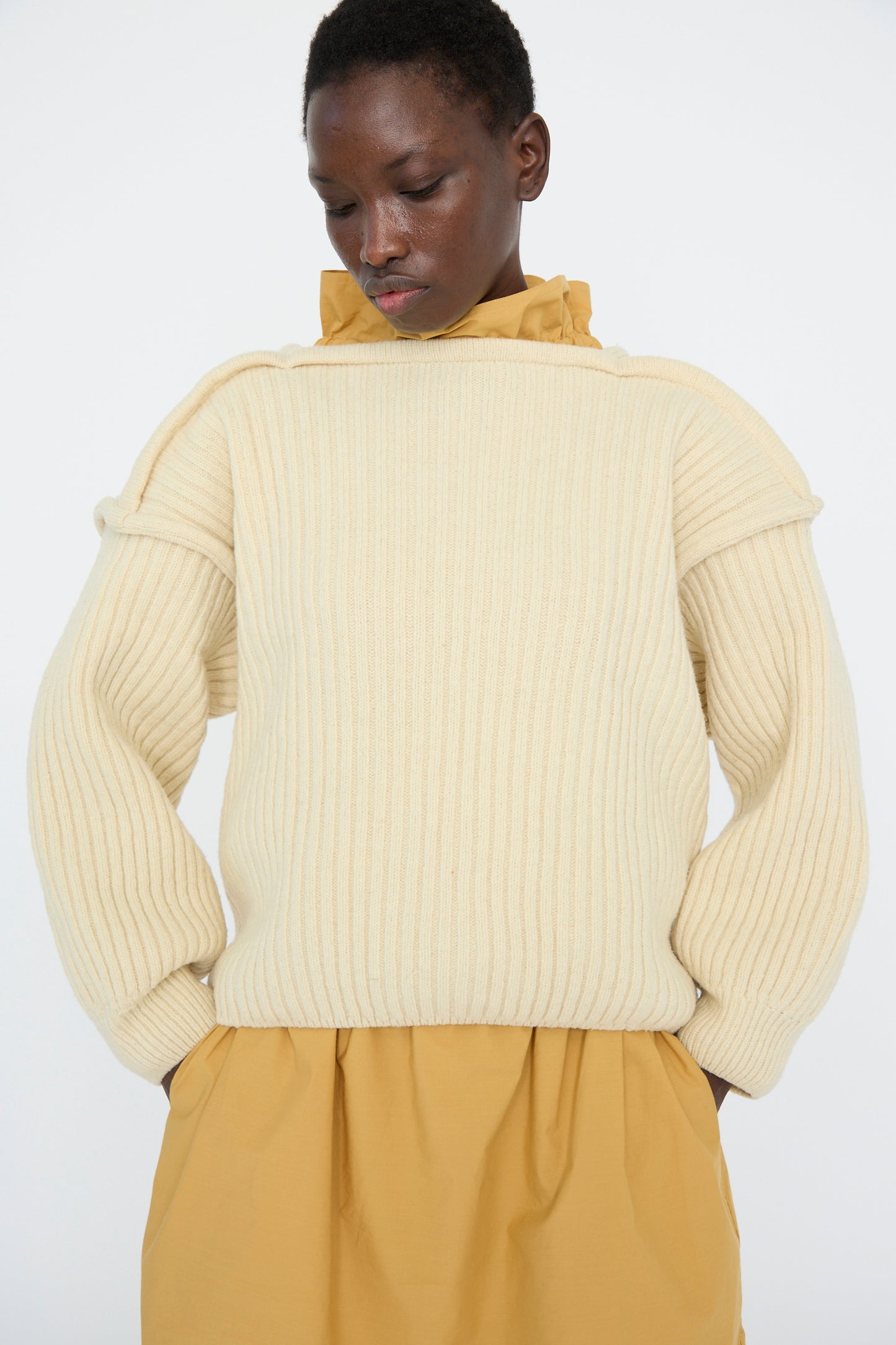 Wearing the Arles Merino Wool Construct Sweater in Ecru by Baserange, the model stands with hands in pockets over a mustard dress against a plain backdrop.