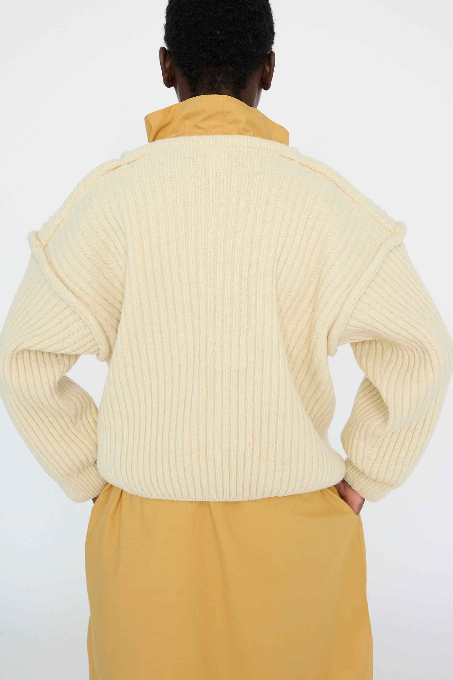 A person faces away from the camera, wearing an oversized Baserange Arles Merino Wool Construct Sweater in Ecru. The sweater's vertical ribbing is styled over a mustard shirt, with hands on hips.