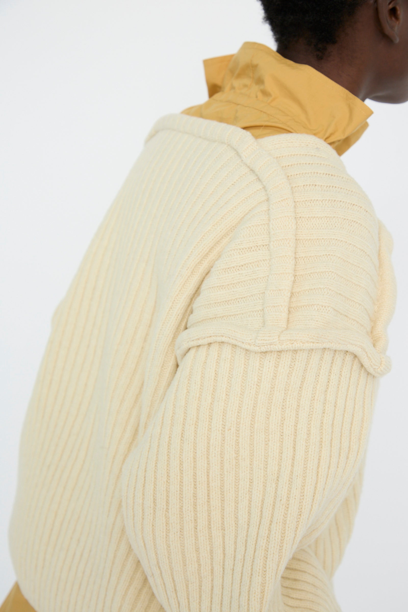 A person faces away wearing the Baserange Arles Merino Wool Construct Sweater in Ecru, featuring vertical ribbing, layered over a mustard yellow collared shirt against a light background.