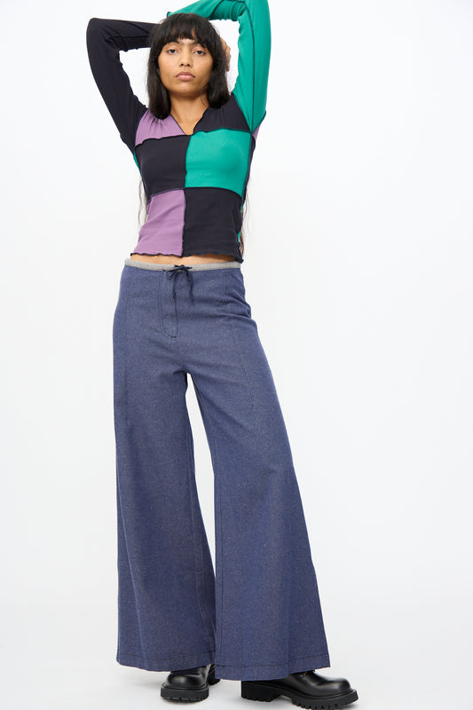 A person stands with arms raised behind their head, wearing a color-blocked long-sleeve shirt, Baserange Arthur Loose Pants in Recycled Indigo, and black shoes against a plain white background.