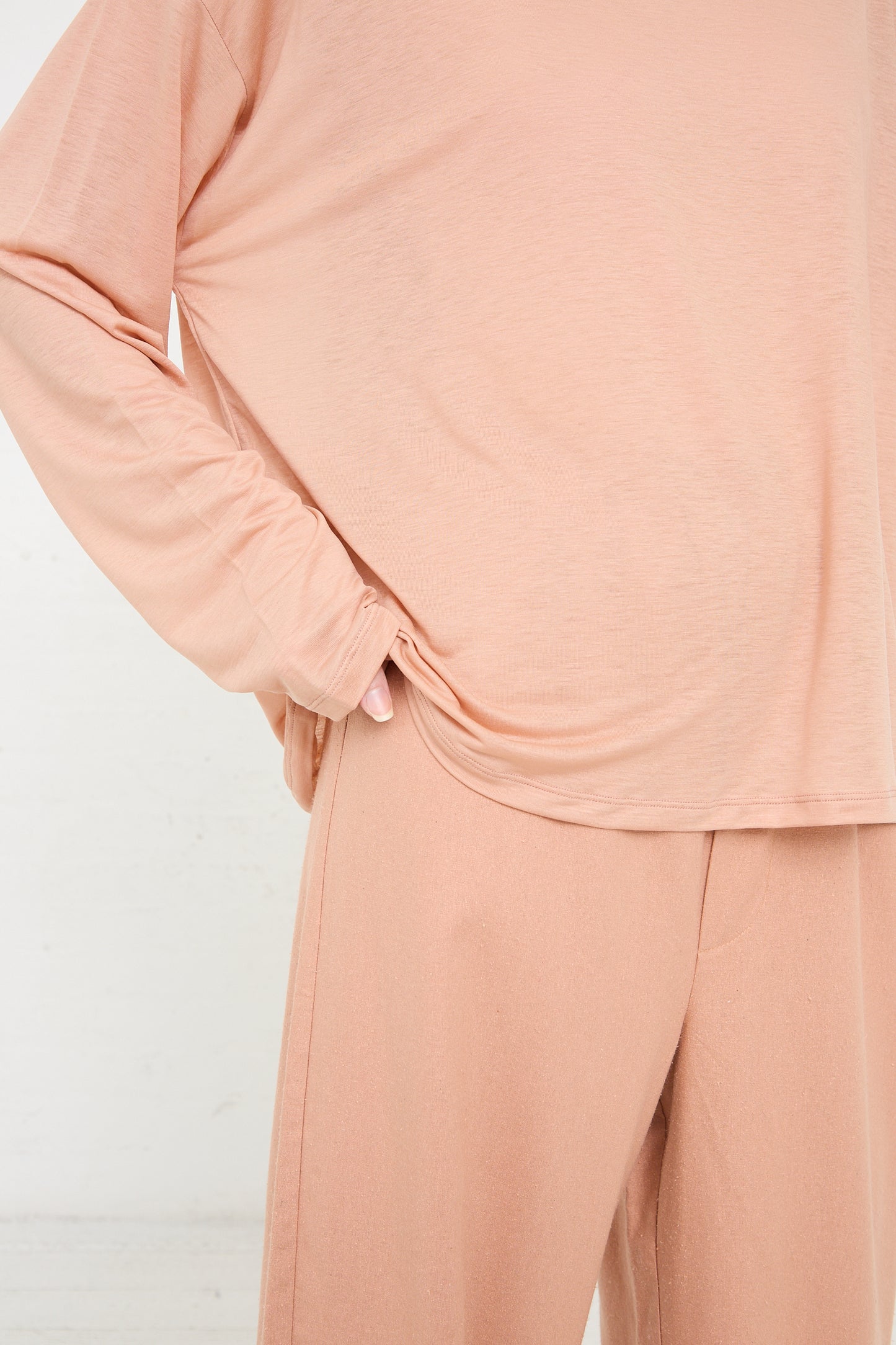Person wearing a Peach-colored Bamboo Loose Long Sleeve Tee in Yu Rose by Baserange with the long sleeve top slightly lifted to reveal the waistband of matching pants, all made from sustainably produced bamboo lyocell.