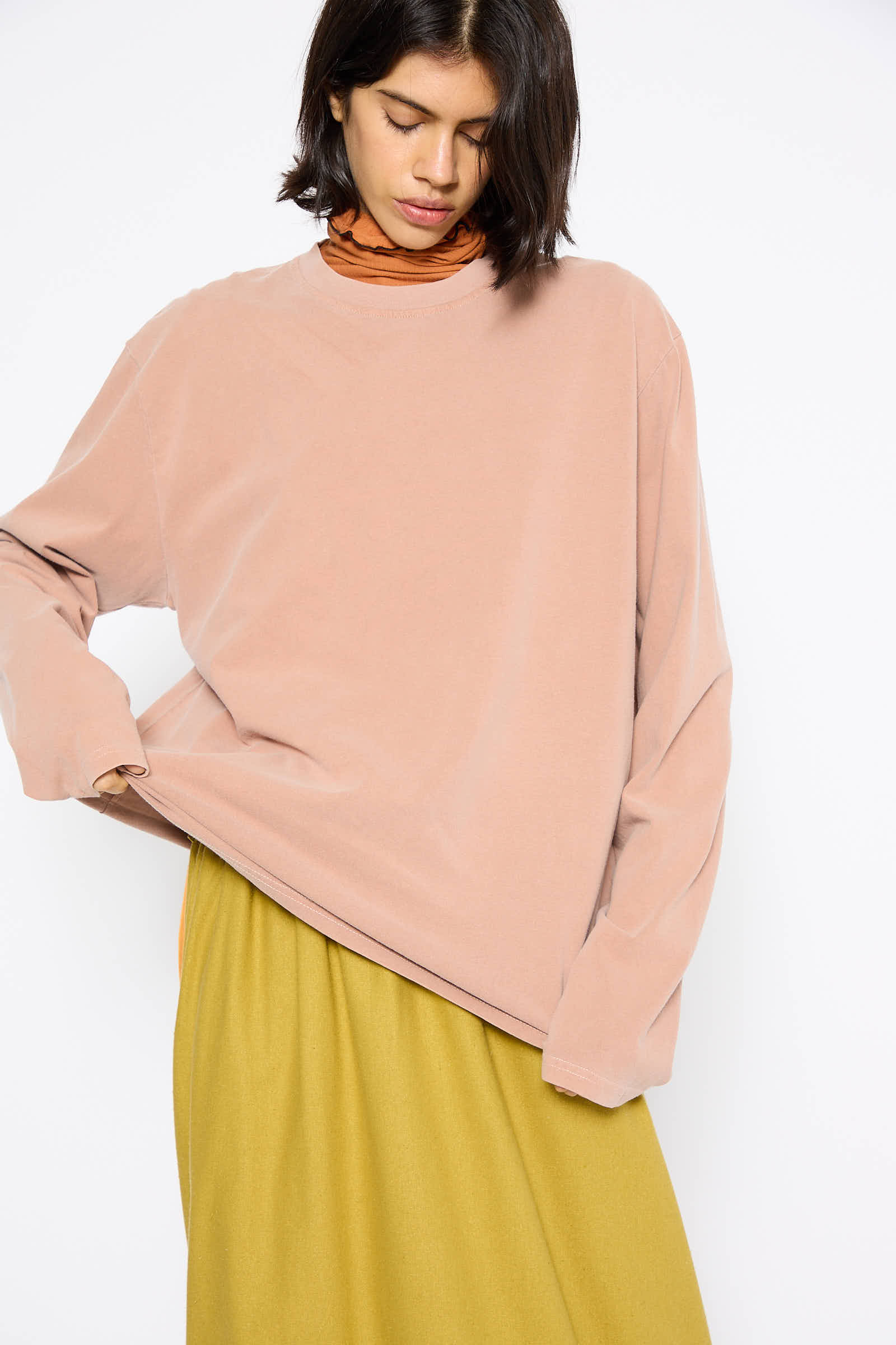 A person wearing the Briar Long Sleeve in Garnet Orange by Baserange, featuring an oversized fit and made from naturally dyed, regenerative cotton, paired with a mustard-yellow skirt, looking downwards against a plain white background.