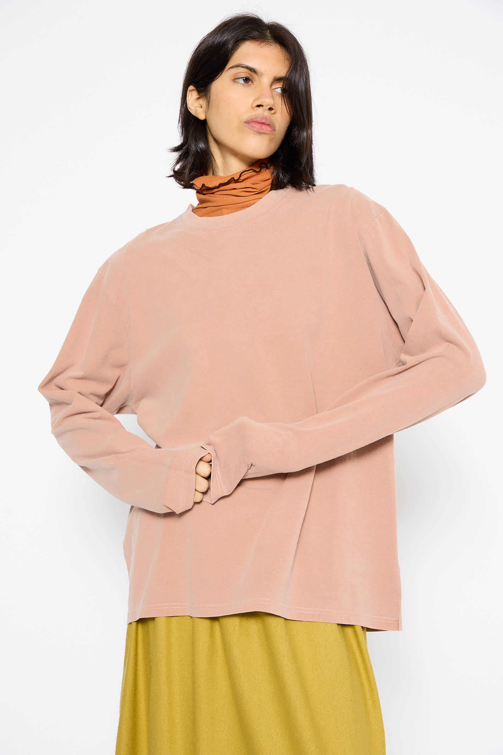 A person with shoulder-length hair wears a light pink oversized sweater, the Briar Long Sleeve in Garnet Orange by Baserange, and a mustard yellow skirt made from naturally dyed fibers, standing against a plain white background.