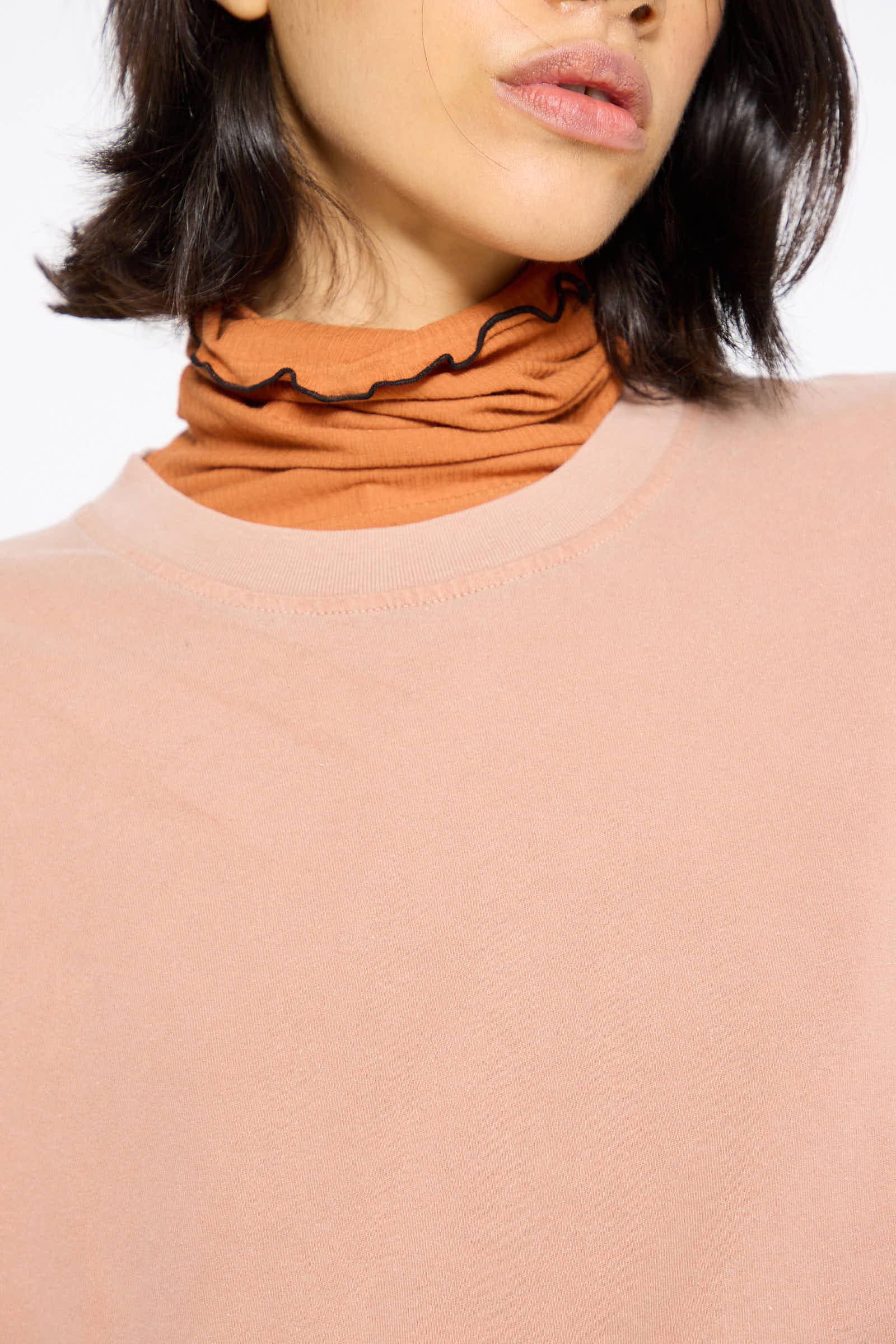 Close-up of a person wearing a naturally dyed pink shirt over the Baserange Briar Long Sleeve in Garnet Orange, which features frilled edges. The relaxed fit complements their short, dark hair, with only the lower half of their face visible.