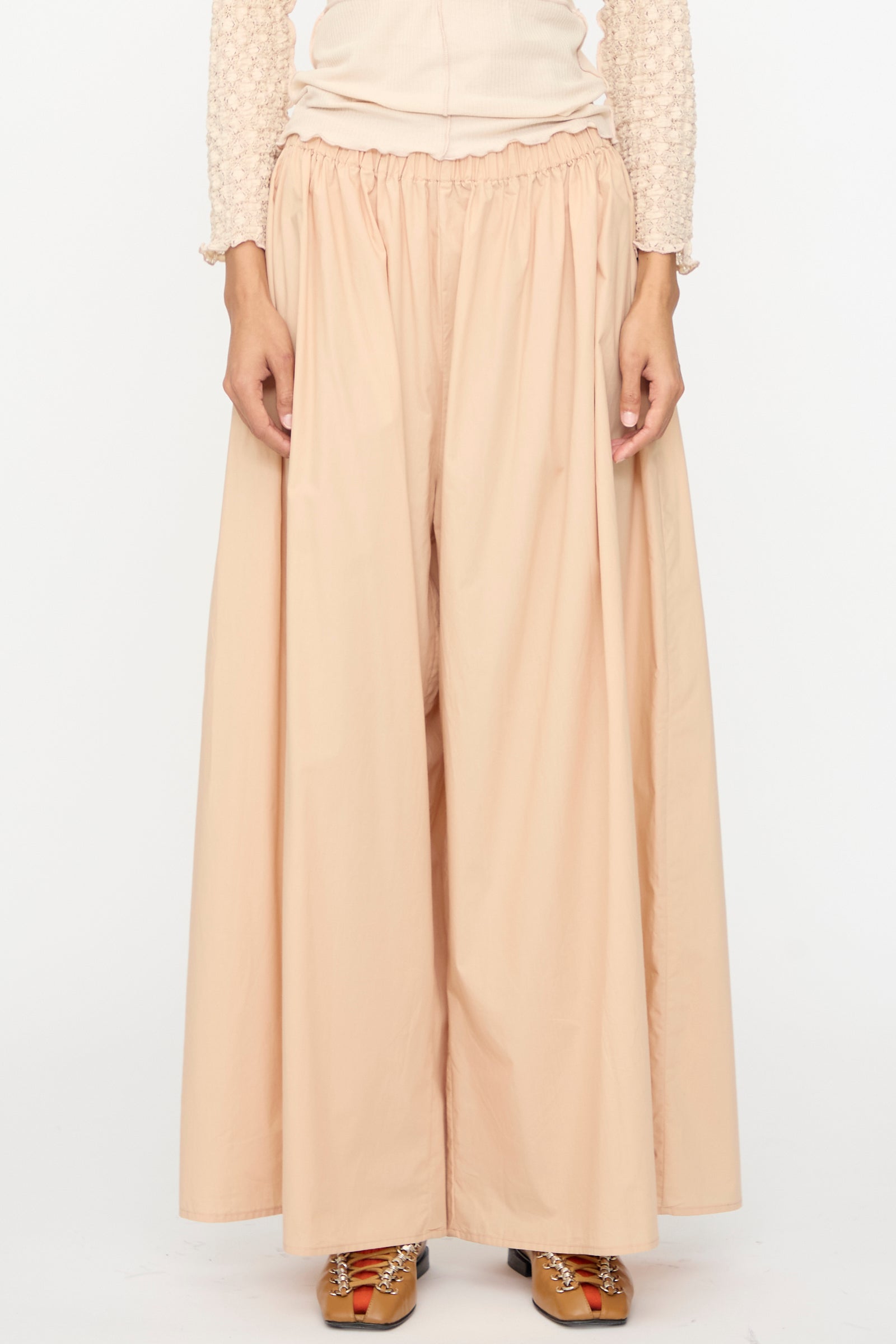 A person wearing Baserange Cable Pants in Andradita Sand with an elastic waist and a light long-sleeve top, standing against a plain white background.