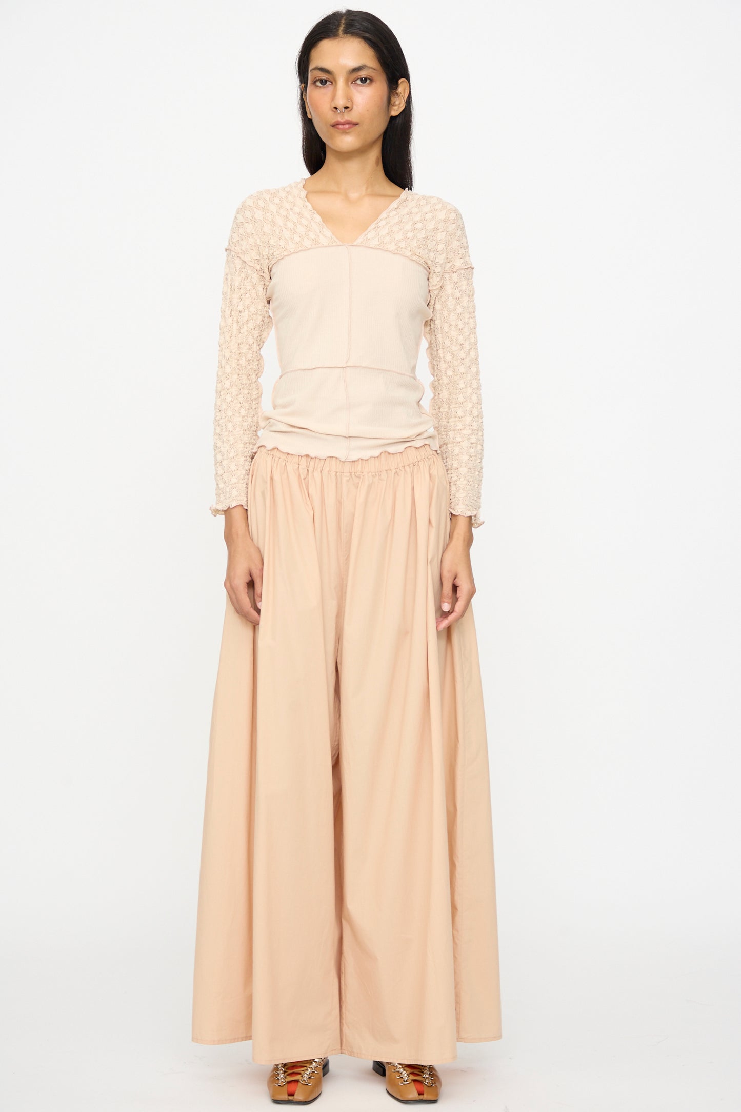 A person stands facing forward in Baserange's Cable Pants in Andradita Sand, featuring an elastic waist, paired with a long-sleeved beige top with patterned sleeves, against a plain white background.