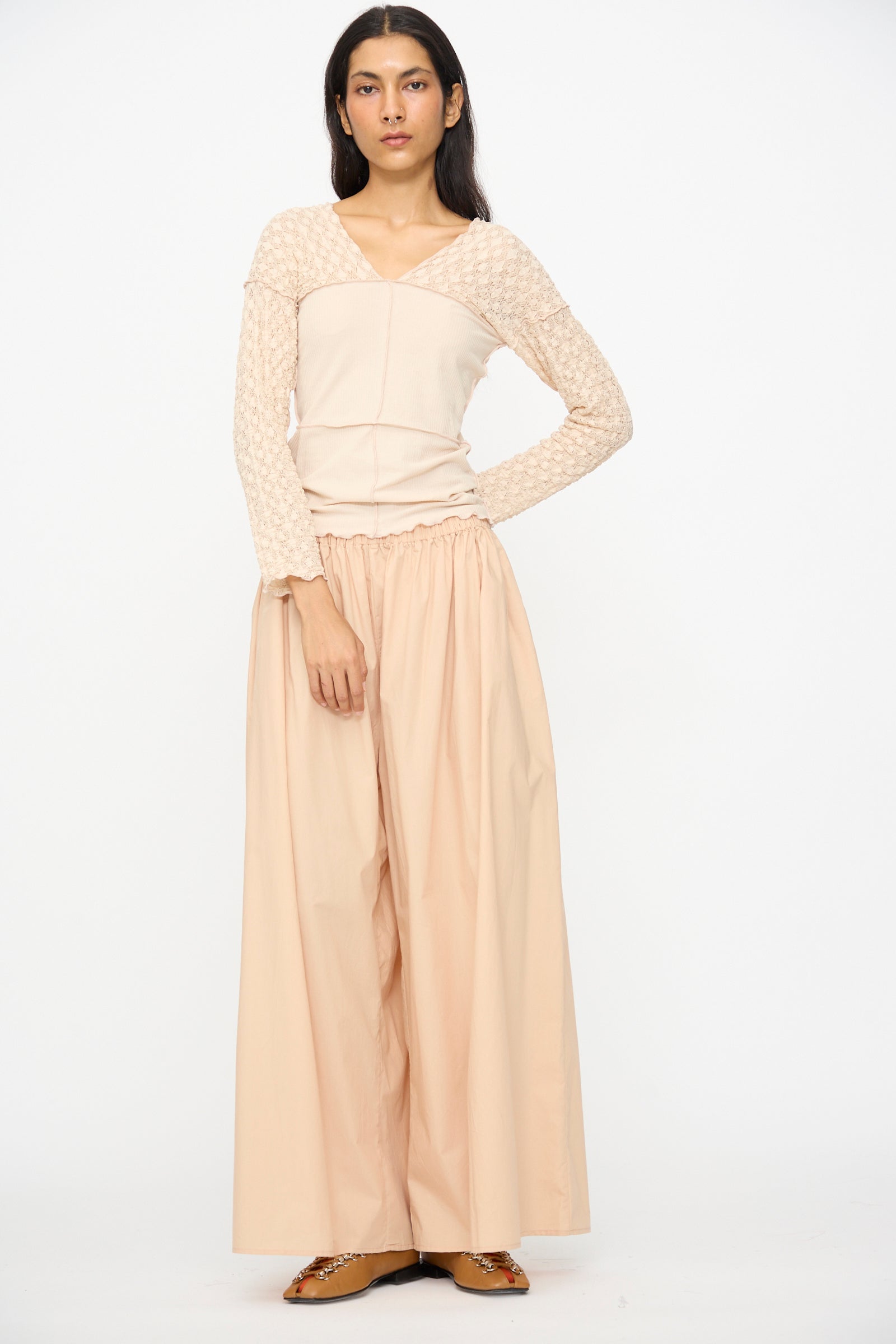 A person stands against a white background wearing Baserange's Cable Pants in Andradita Sand with an elastic waist, paired with a light beige long-sleeve top and brown flat shoes.