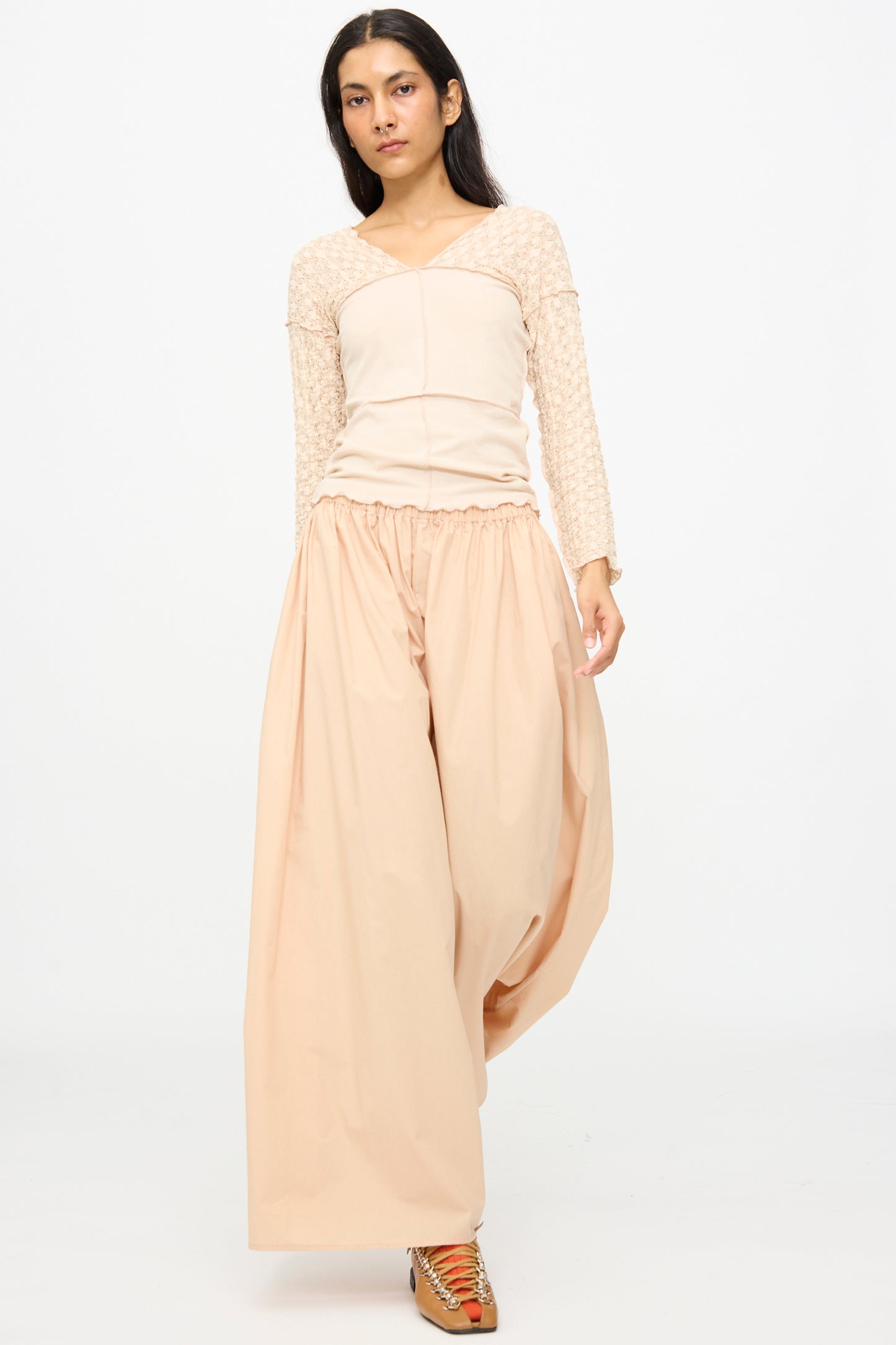 A person walks forward against a plain white background wearing a beige long-sleeve top and Baserange's Cable Pants in Andradita Sand, featuring loose, wide cotton legs and an elastic waist.