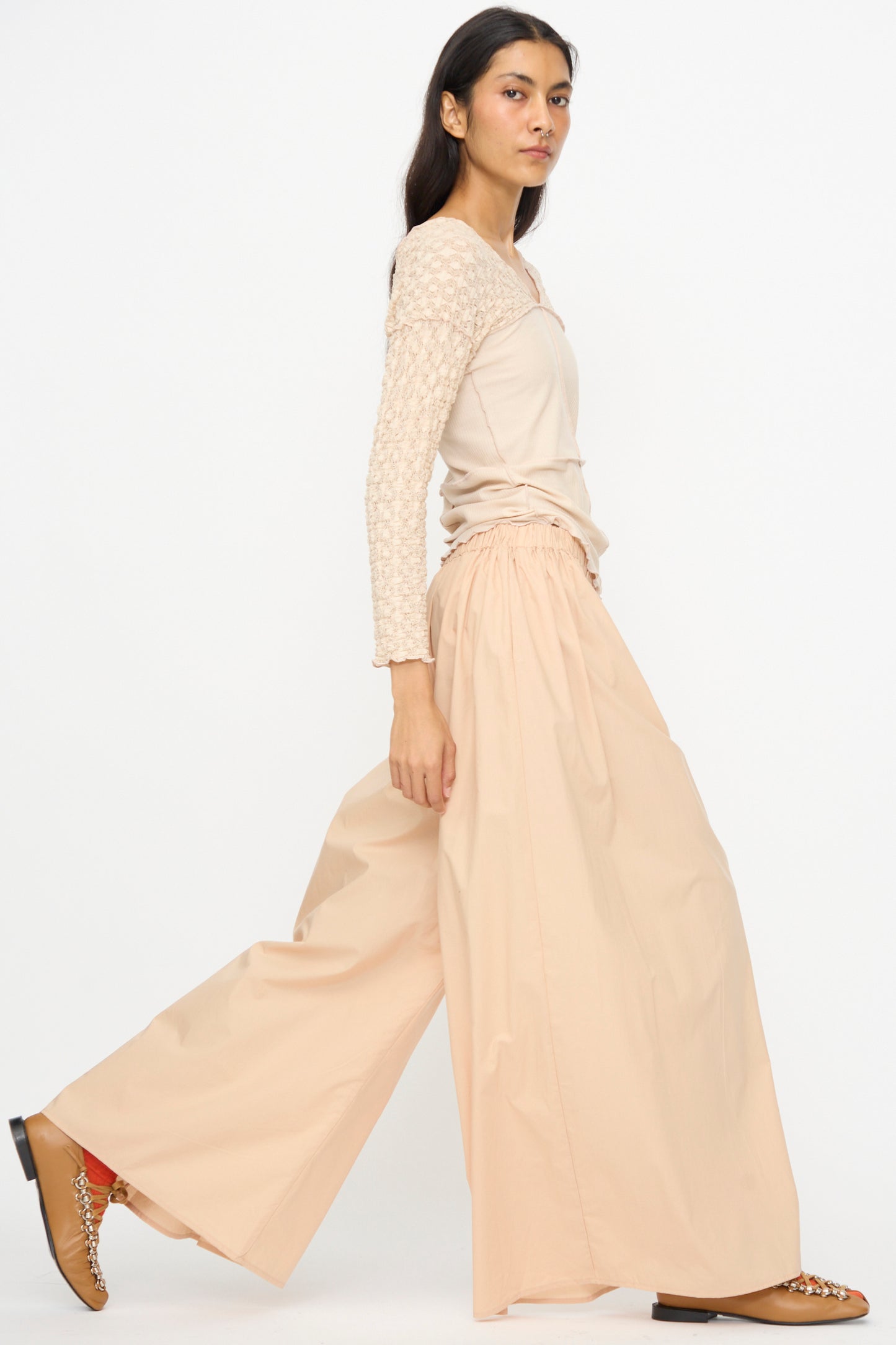 A woman wears Baserange's Cable Pants in Andradita Sand—wide cotton pants with an elastic waist—and a light beige, long-sleeve cotton poplin top. She has on brown shoes with chain details and stands against a plain white background.