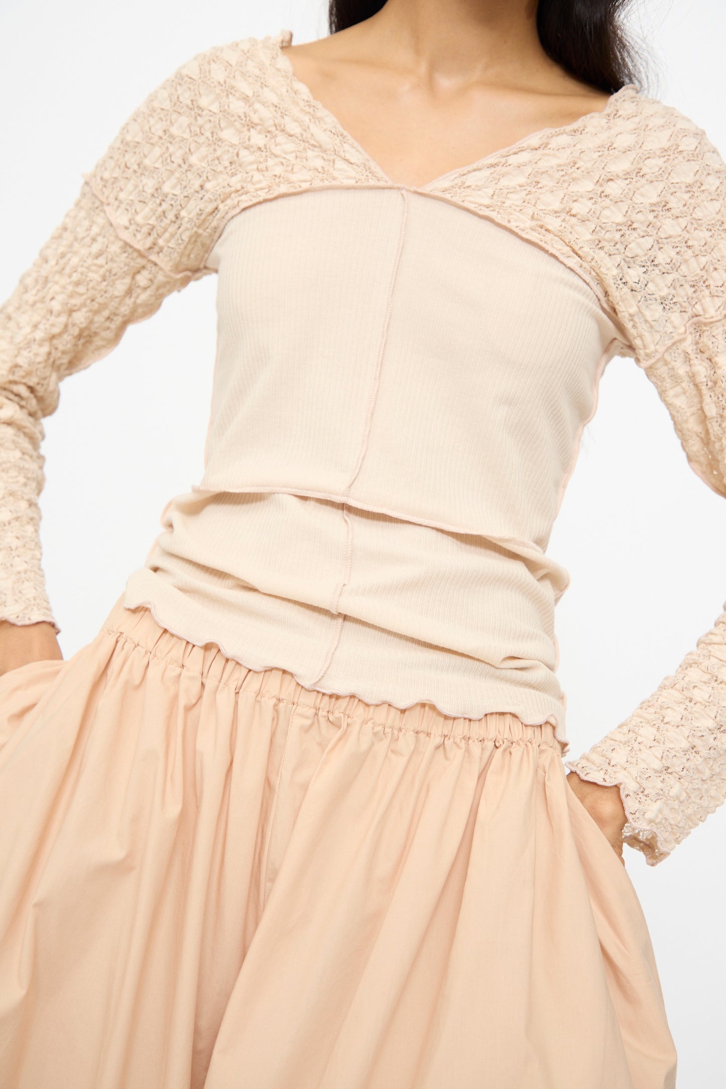 A person is wearing Baserange’s Cable Pants in Andradita Sand. Their cream, long-sleeve lace top has a textured bodice. The face is partially out of frame.