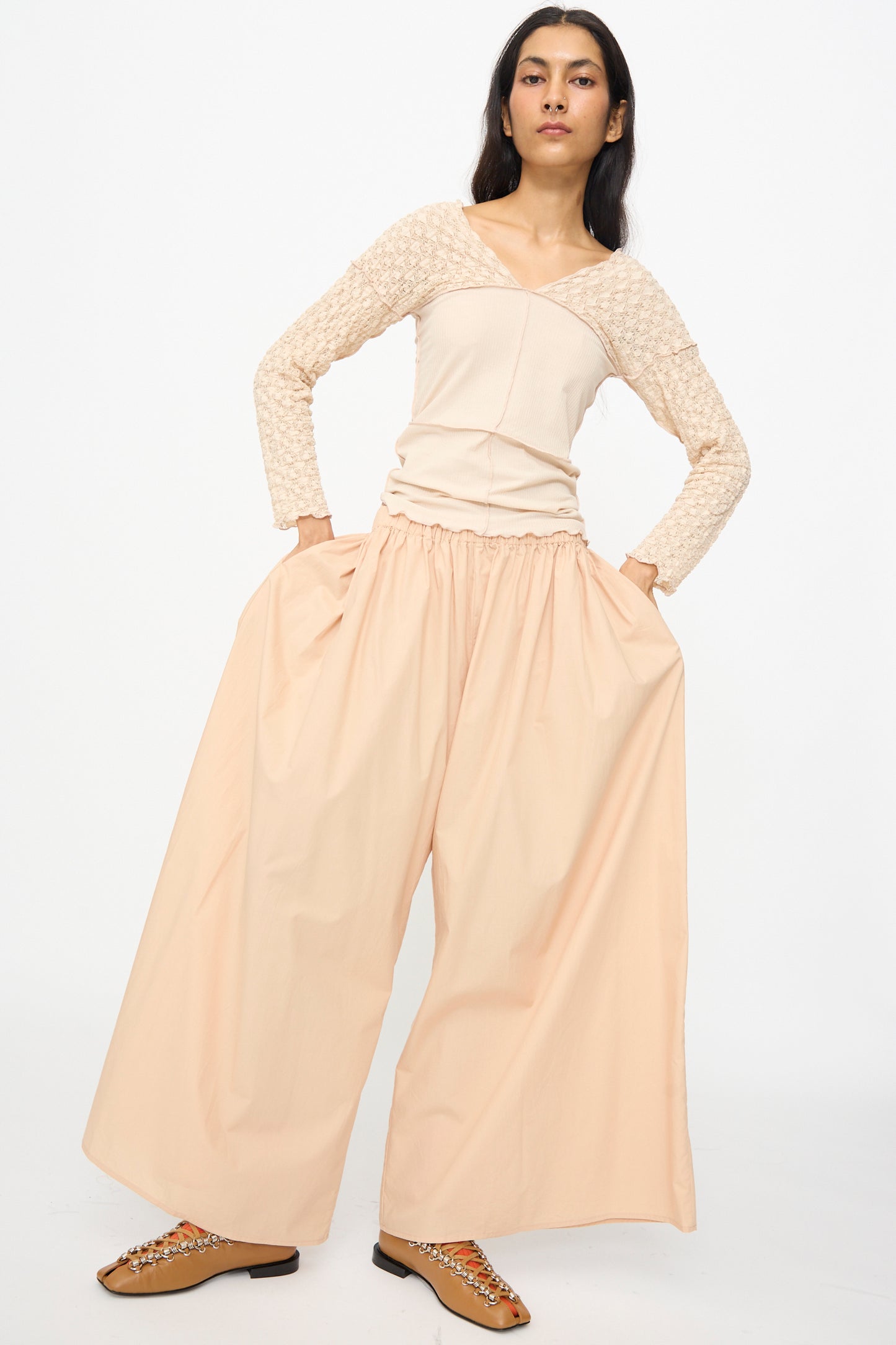 A person stands with hands in pockets, wearing Baserange Cable Pants in Andradita Sand—wide cotton pants with an elastic waist—paired with a textured long-sleeve top and embellished brown flats against a plain white background.